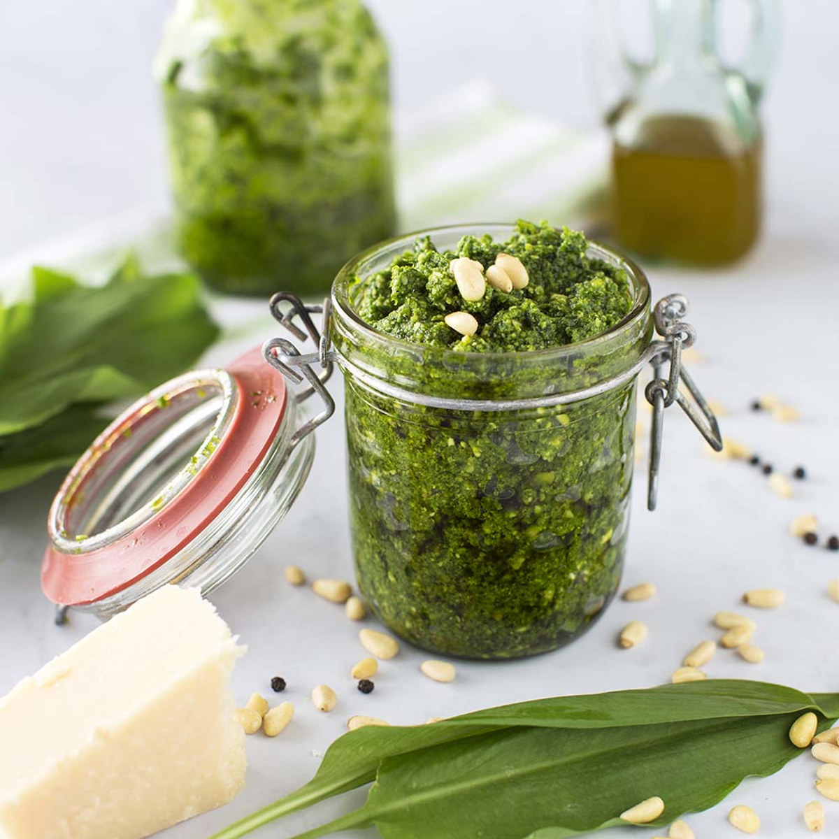 Słoik pesto z orzeszkami piniowymi na wierzchu, otoczony składnikami i kawałkiem sera.