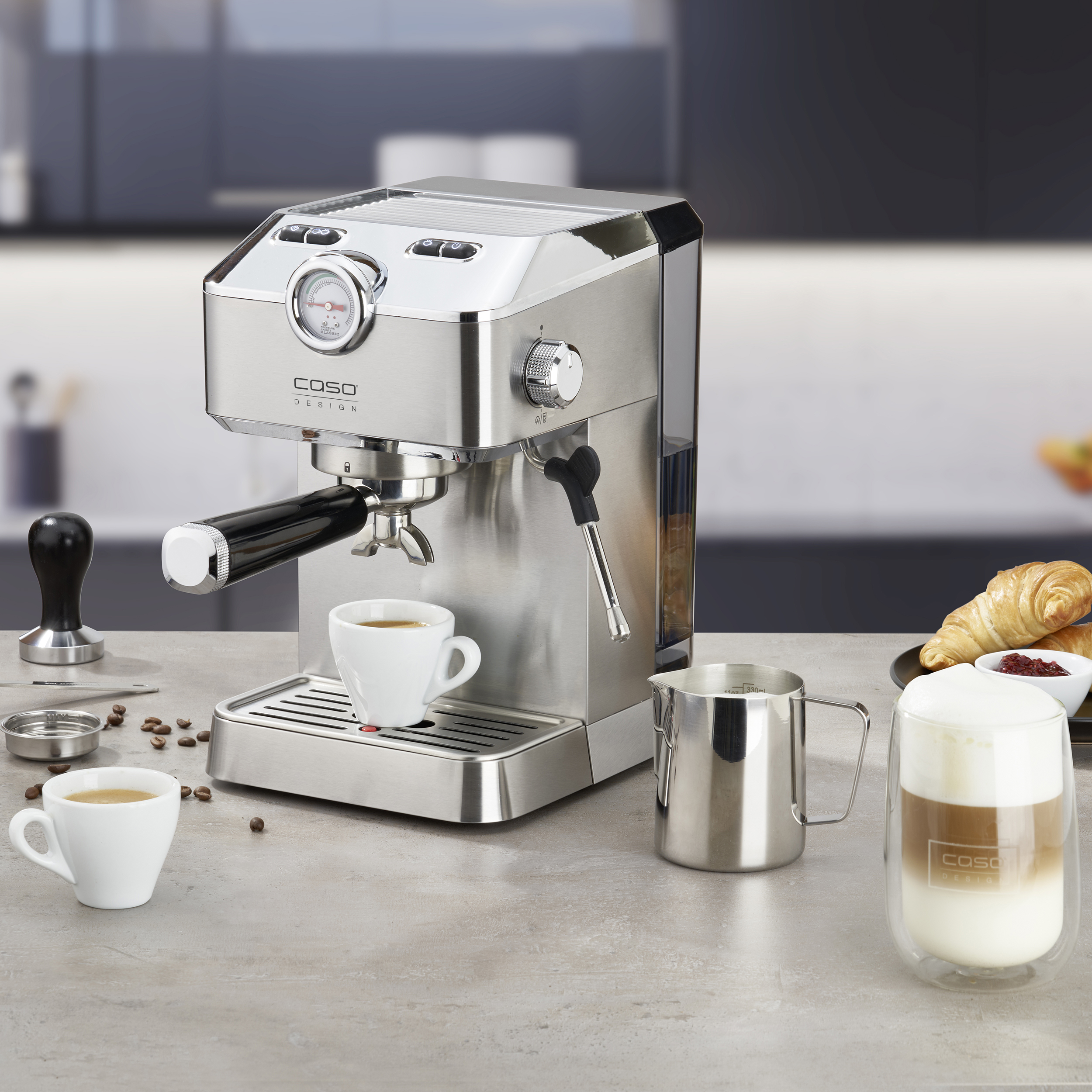 Eine Espressomaschine aus Edelstahl mit Tassen, Kaffee und einem Milchschaumkännchen auf einer Arbeitsplatte.