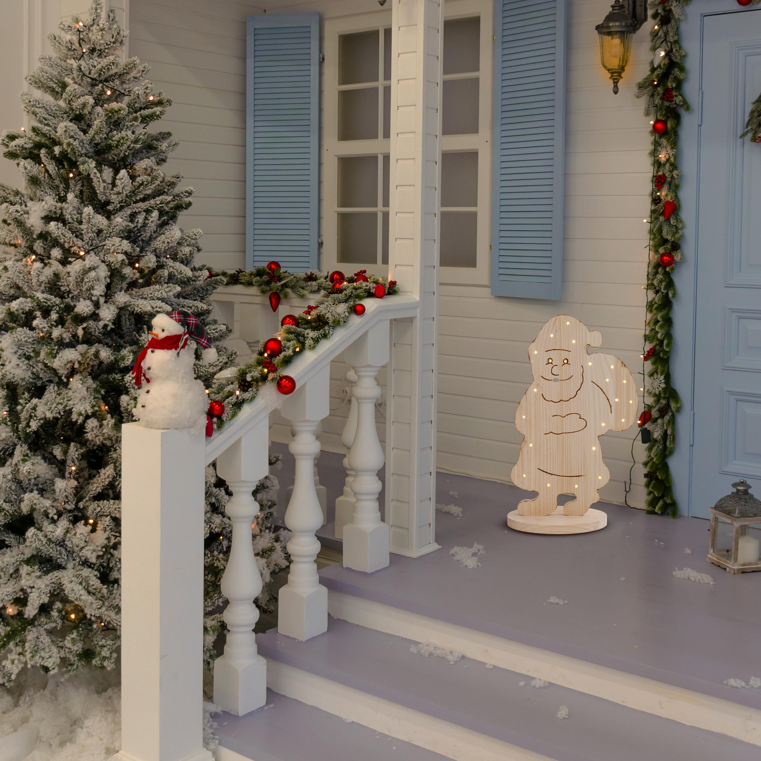 Weihnachtsszene mit Schneemann und hölzernem Weihnachtsmann auf einer Veranda. Girlande mit roten Kugeln und einem schneebedeckten Baum.