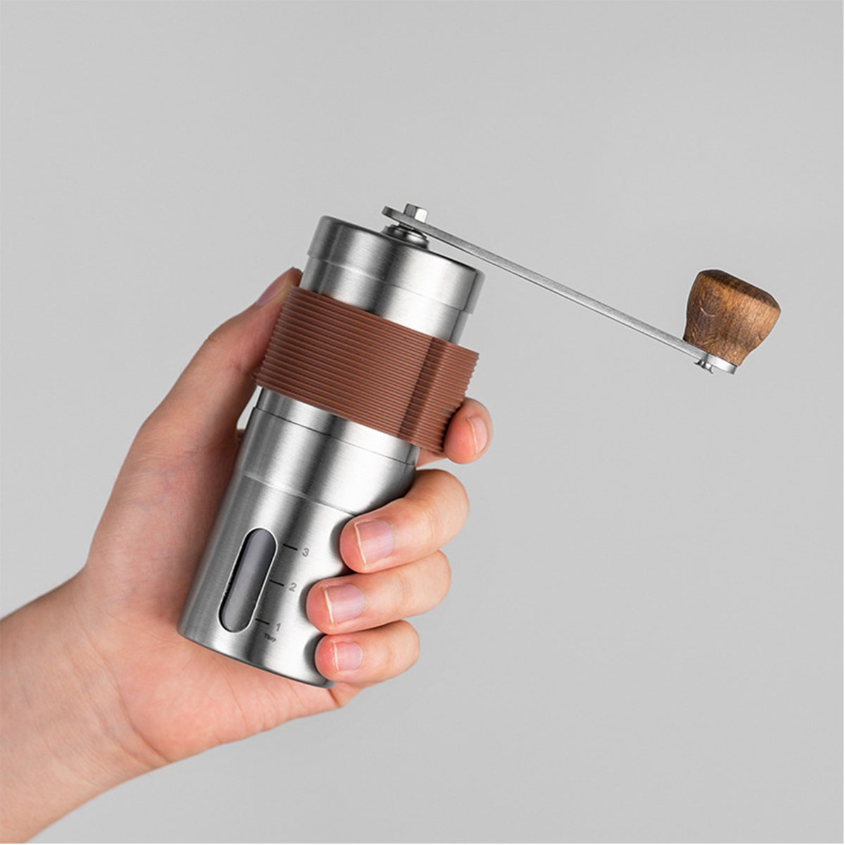 Eine Hand hält eine silberne manuelle Kaffeemühle mit braunem Griff vor grauem Hintergrund.