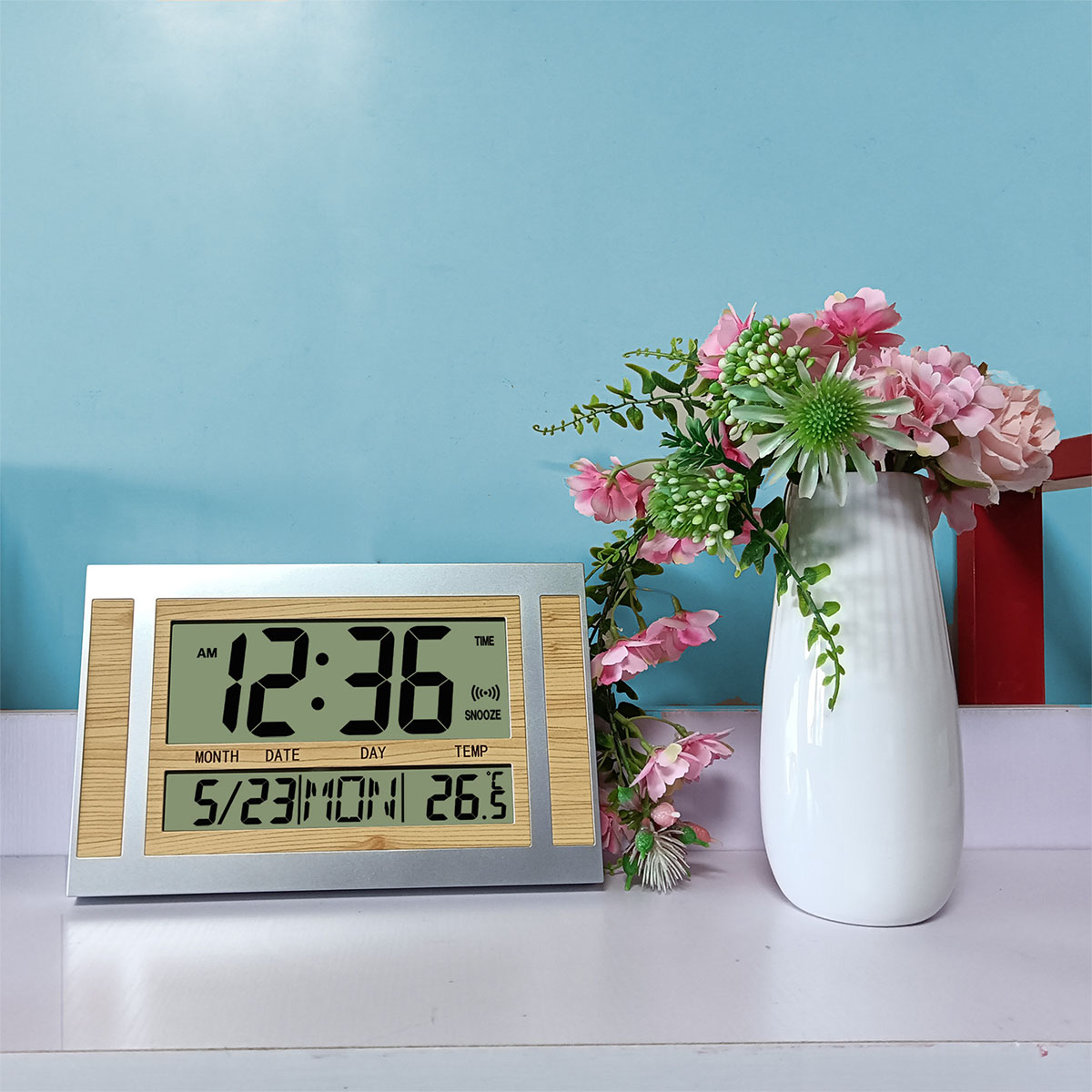 Digitaluhr mit Zeit, Datum und Temperatur. Vase mit rosa Blumen auf weißer Oberfläche vor blauem Hintergrund.