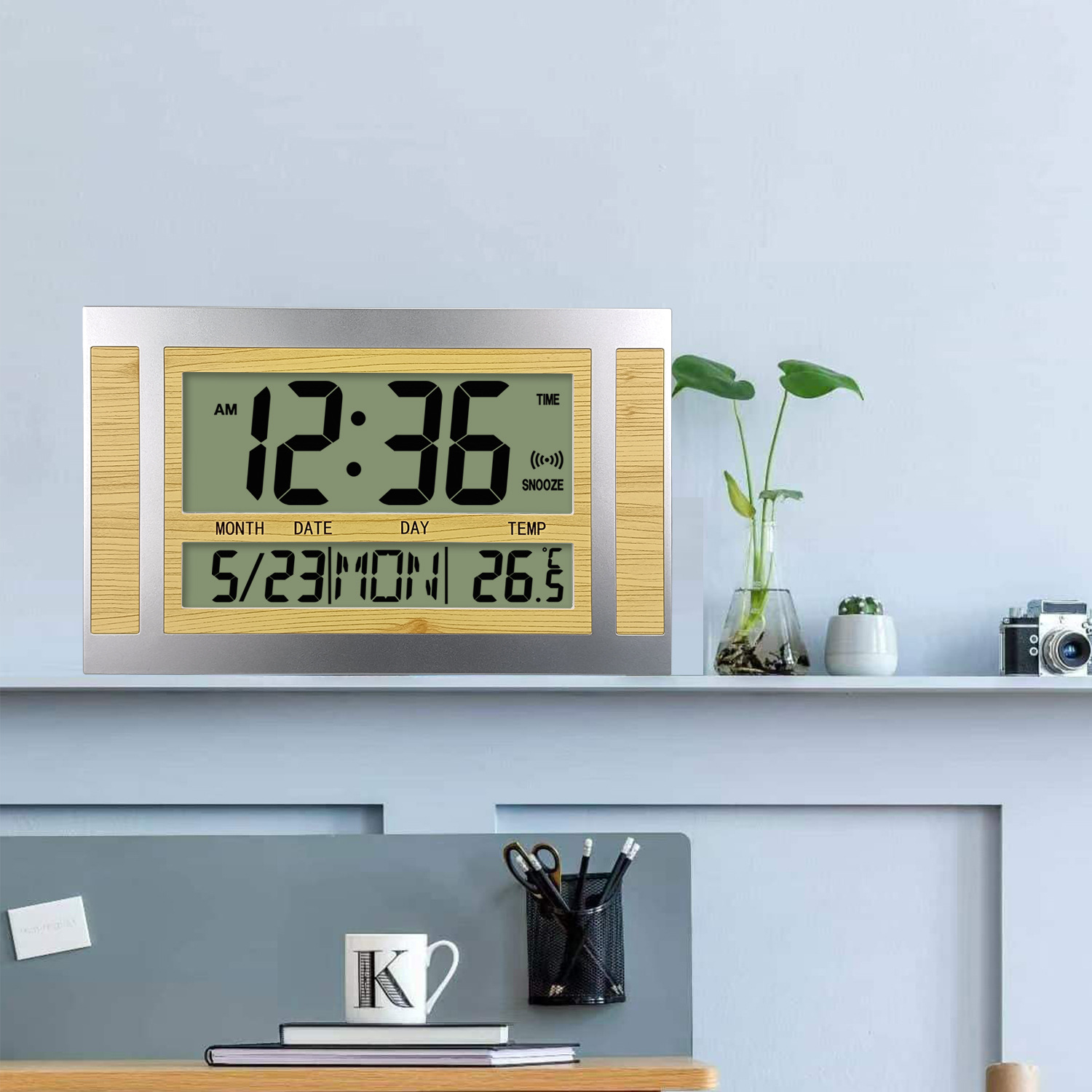 Digitaluhr mit Uhrzeit und Datum. Auf einem Regal mit Pflanze und anderen Objekten.