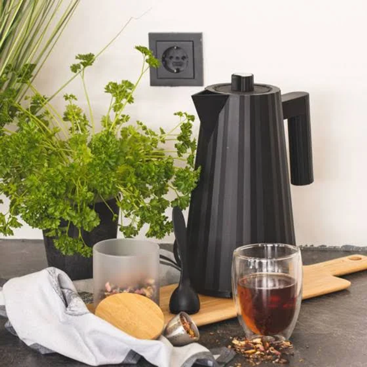 Hervidor negro en tabla de madera. Té, vaso, cuchara y hierbas, en encimera. Pared y enchufe negros.