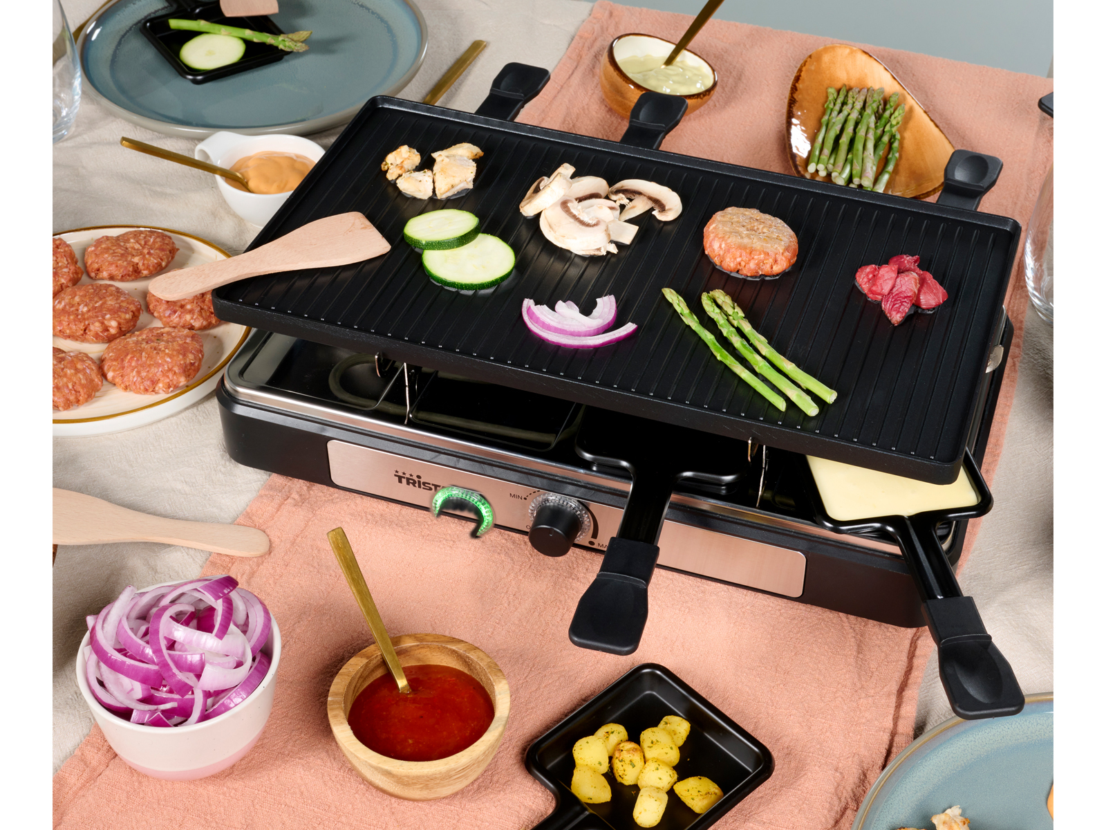 Grill per raclette con cibo tra cui verdure, formaggio e carne, e piccole ciotole.