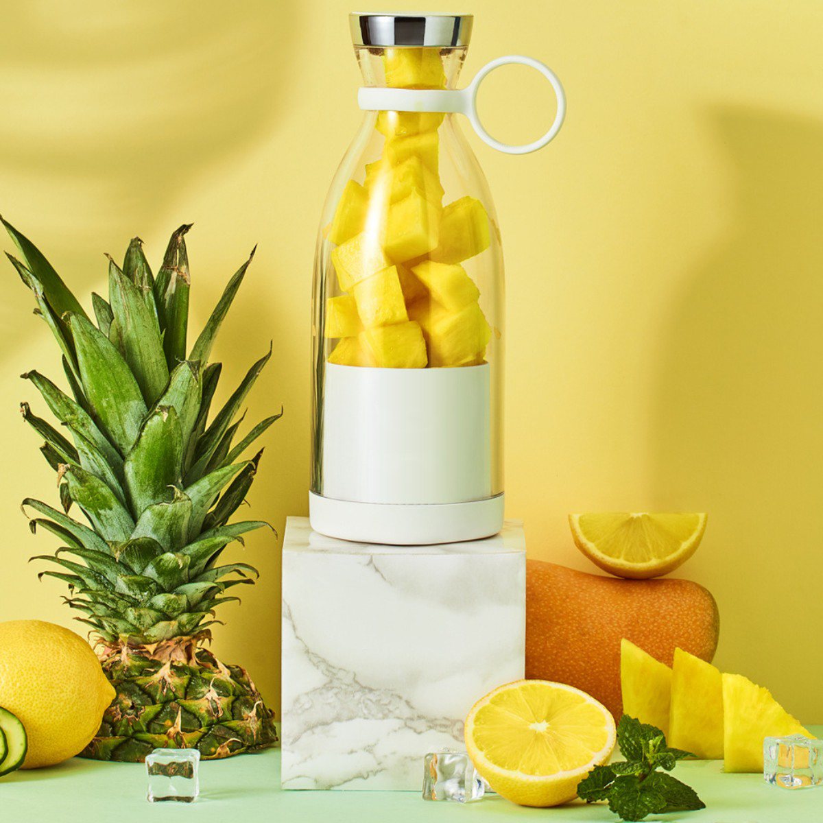 Mixer mit Mango, Zitrone, Ananas und Minze auf weißem Block und gelbem Hintergrund.