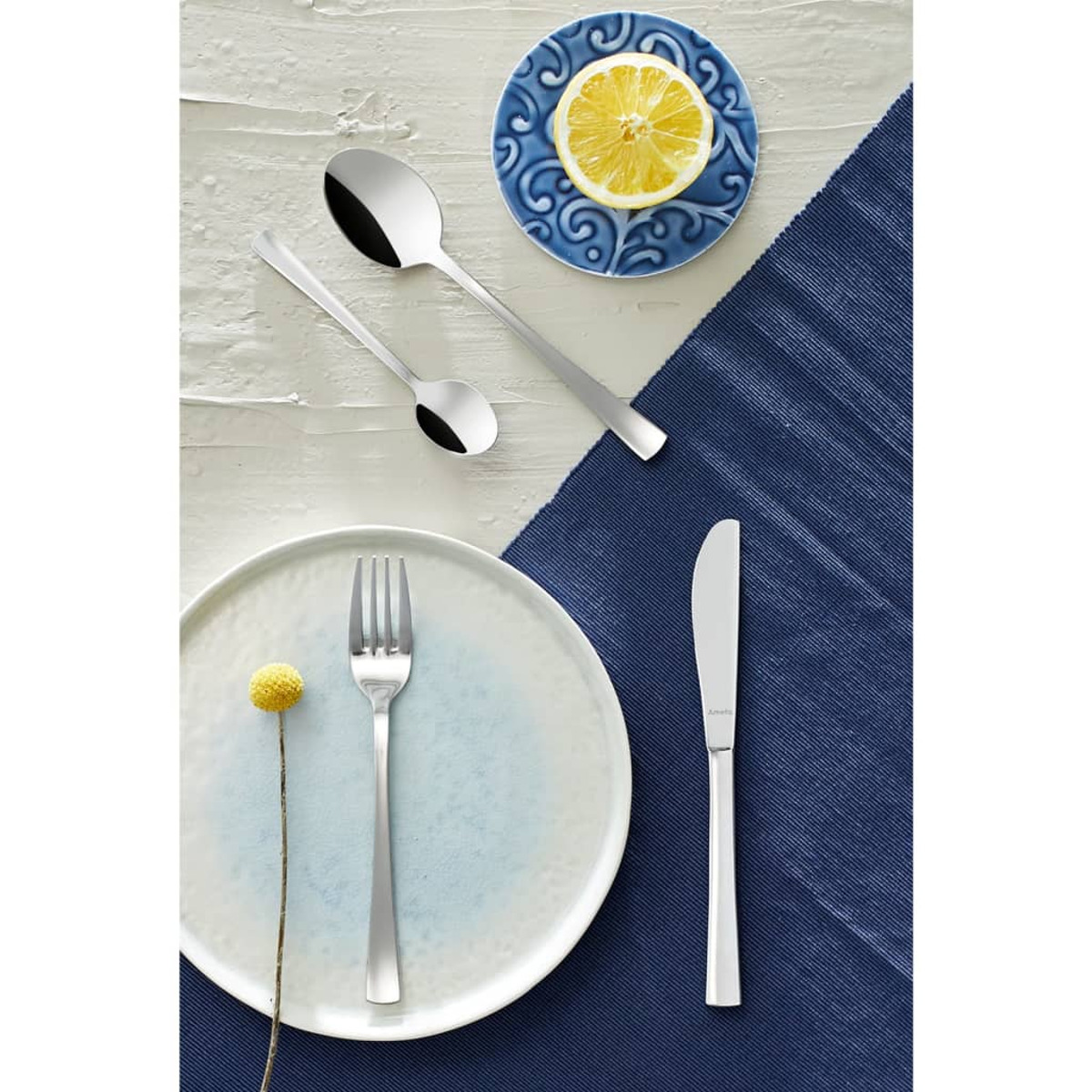 Besteck-Set: Löffel, Gabel und Messer auf einem Tisch mit blauem Platzset und Zitrone auf kleinem Teller.