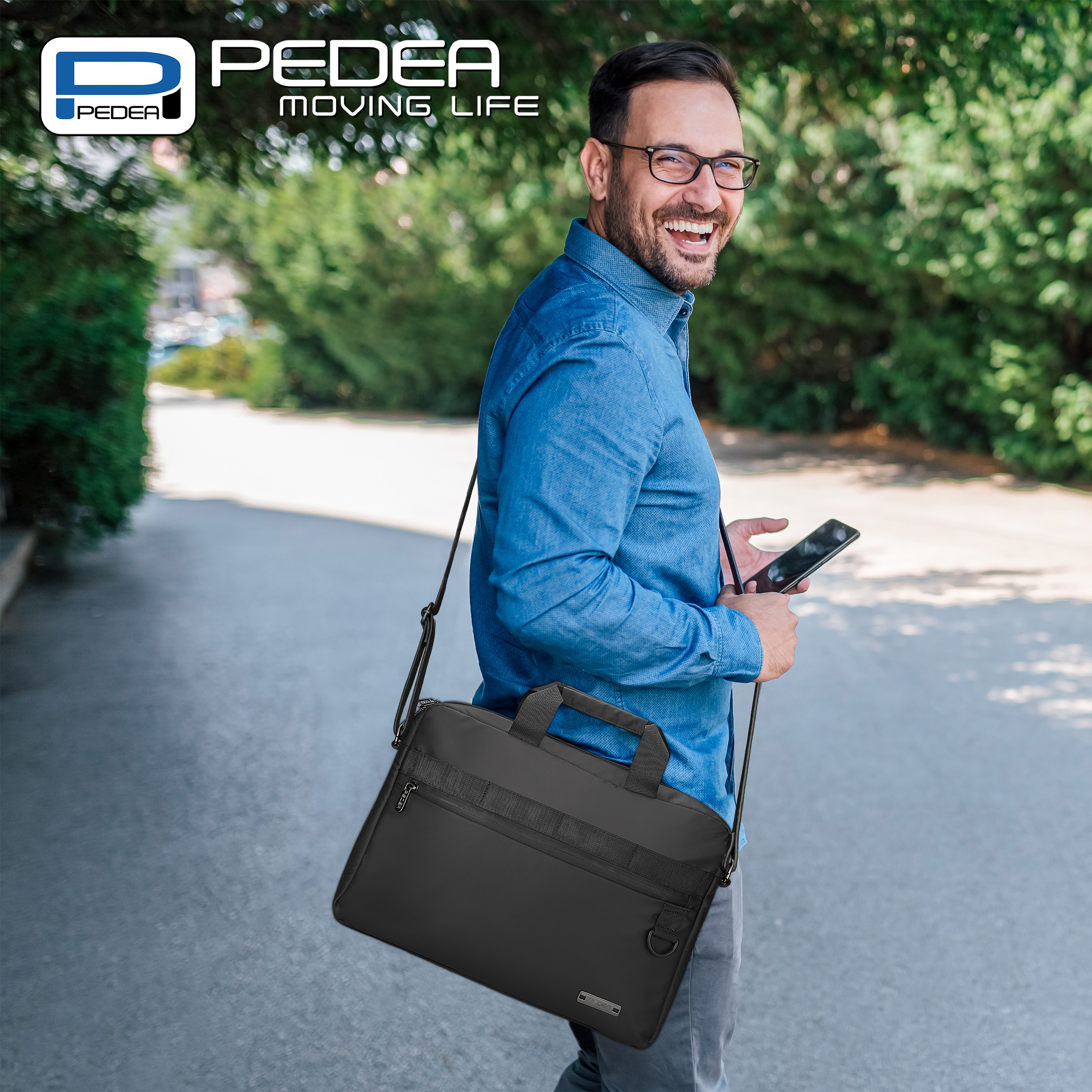 Ein Mann in blauem Hemd mit schwarzer Tasche geht lächelnd auf einem Pfad und hält ein Telefon.