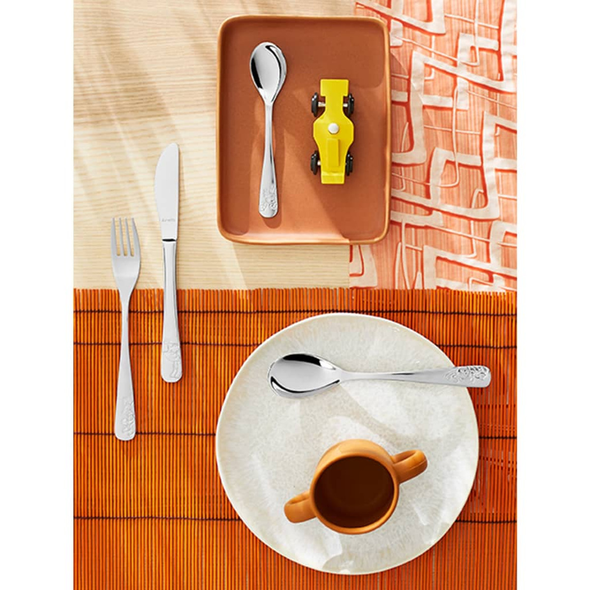 Eine Tischdecke mit Silberwaren, einem gelben Spielzeugauto auf einem Tablett und einer kleinen orangenen Tasse.