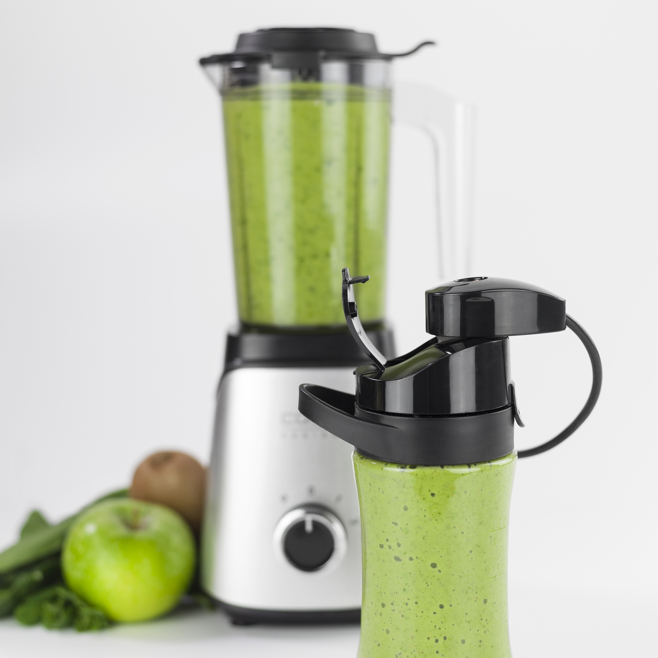 Ein Mixer mit grünem Smoothie und eine gefüllte Flasche vor einem grünen Apfel, Kiwi und Petersilie.
