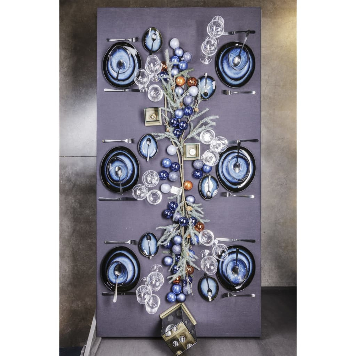 Tischdekoration mit blauen Tellern, Gläsern und Ornamenten. Mittelstück mit Zweigen und Kugeln. Dunkelgrauer Hintergrund.
