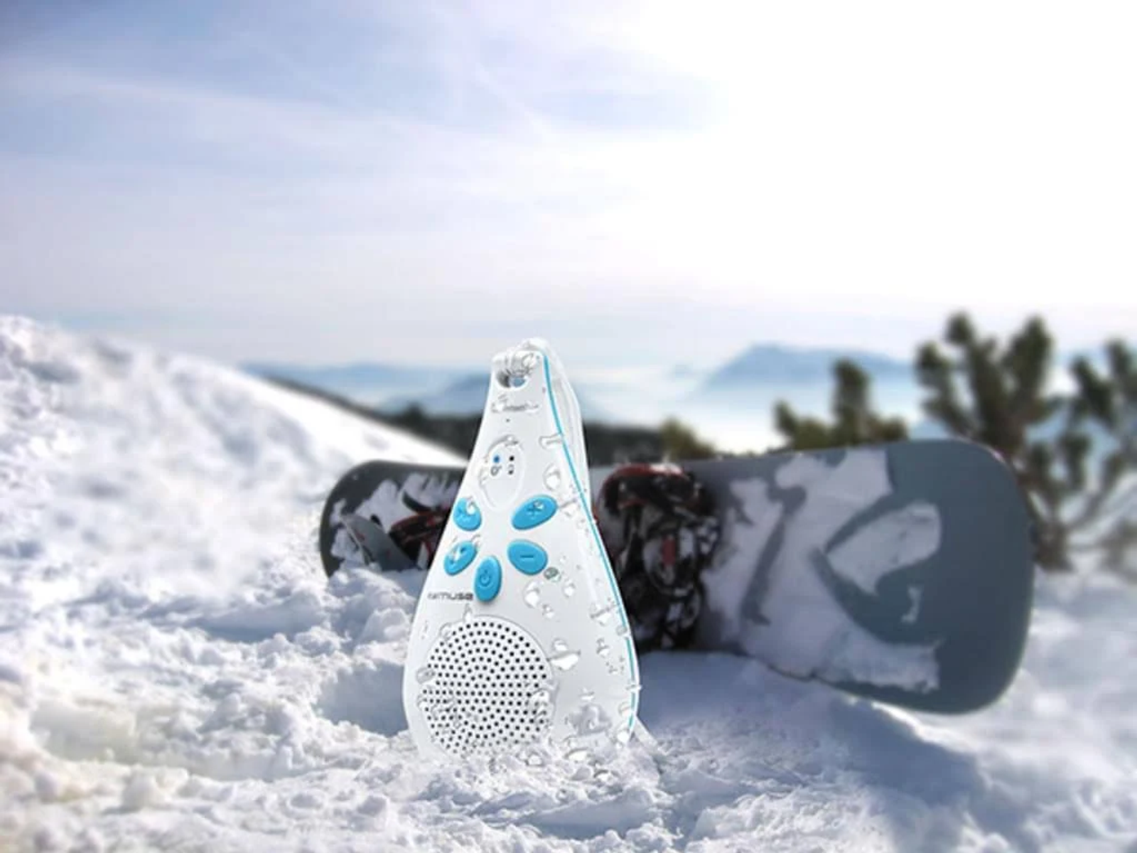 Weiß-blauer Lautsprecher im Schnee, neben einem Snowboard. Verschneiter Hintergrund.