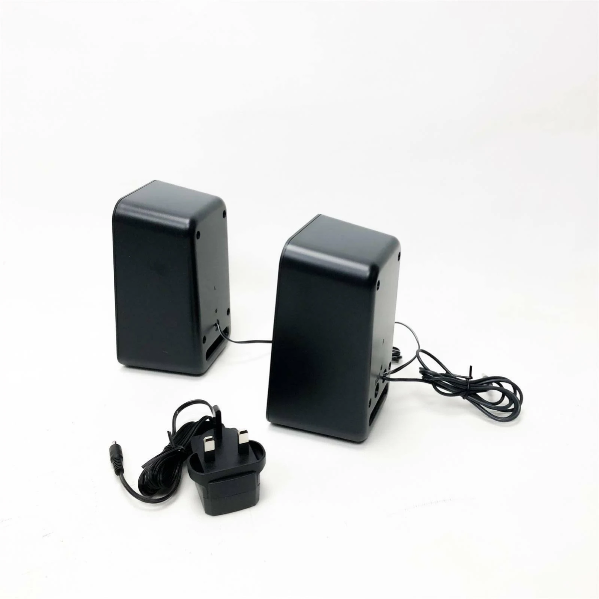 Dos altavoces negros con un adaptador de corriente y cables sobre una superficie blanca.