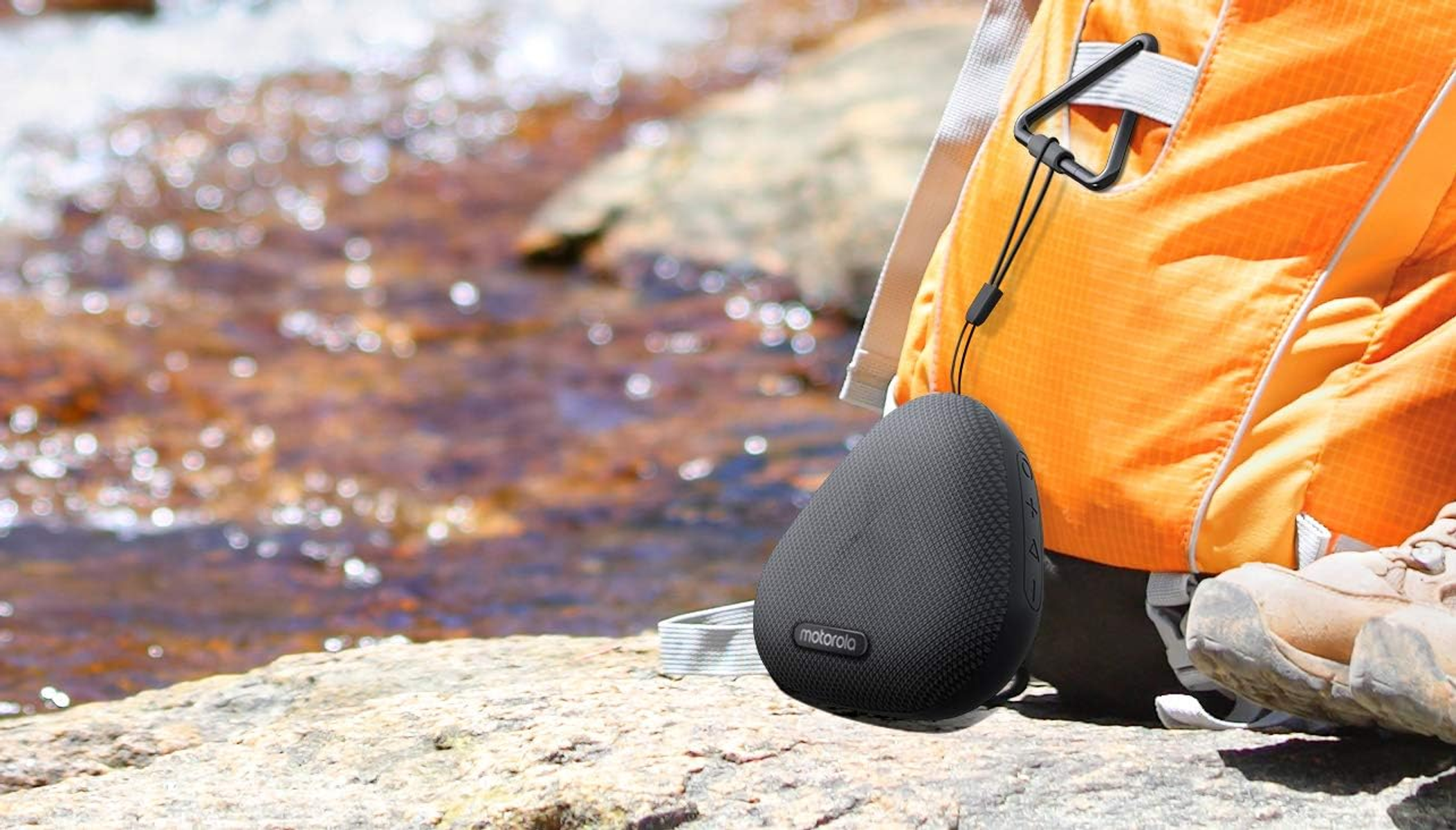 Altavoz Motorola negro sobre una roca junto a un arroyo, junto a una mochila naranja.