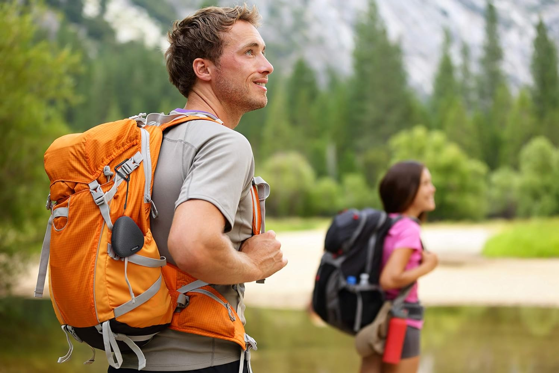 Dos excursionistas con mochilas, una naranja, en un bosque cerca de un río.