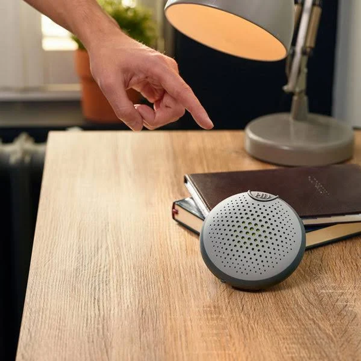 Osoba wskazująca na mały, okrągły, szary głośnik na drewnianym stole. Obecne są książki i lampa.