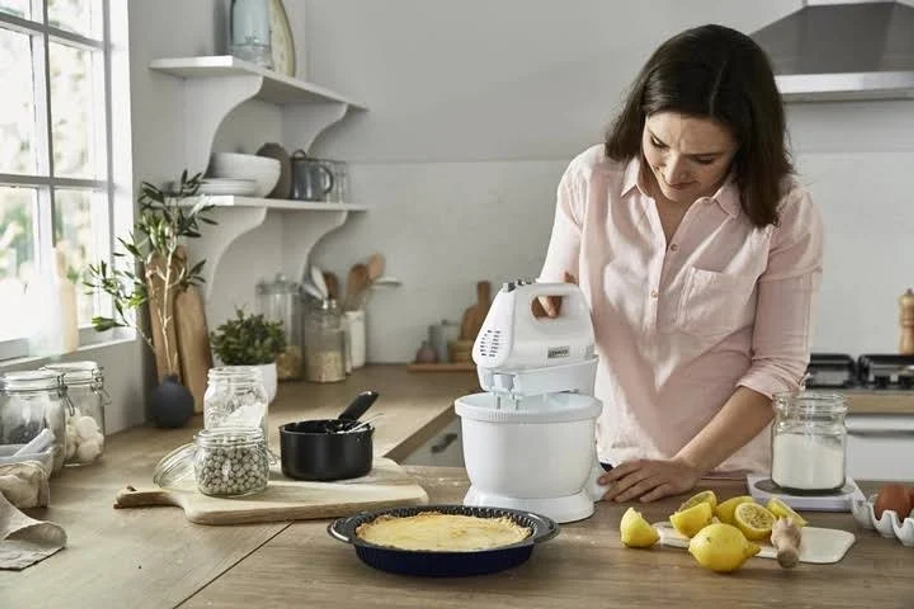 Una donna usa un mixer in cucina. Ci sono ingredienti e una torta sullo sfondo.