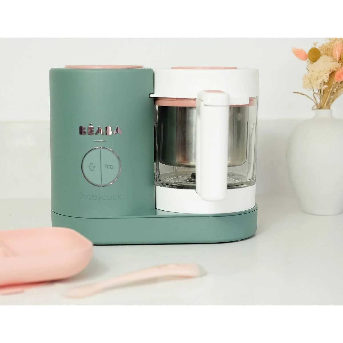Robot da cucina per neonati verde e bianco con dettagli rosa e accessori per neonati, su una superficie bianca.