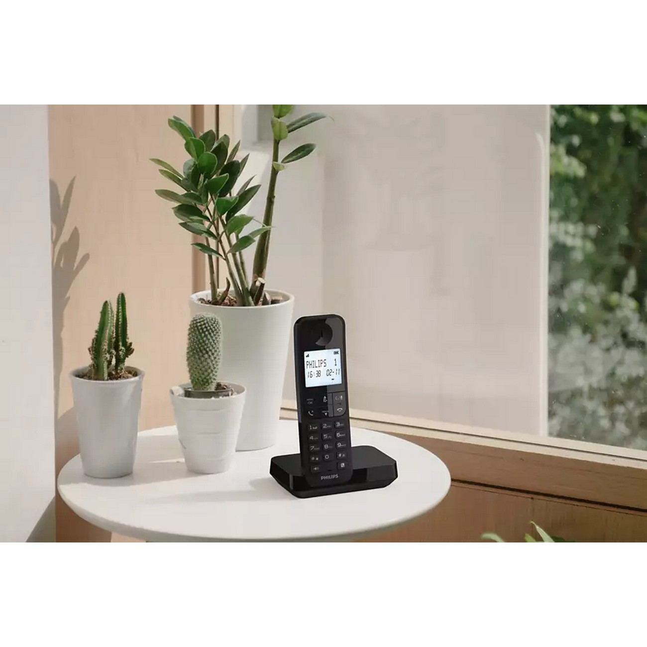 Teléfono inalámbrico en una estación base junto a plantas y una ventana.