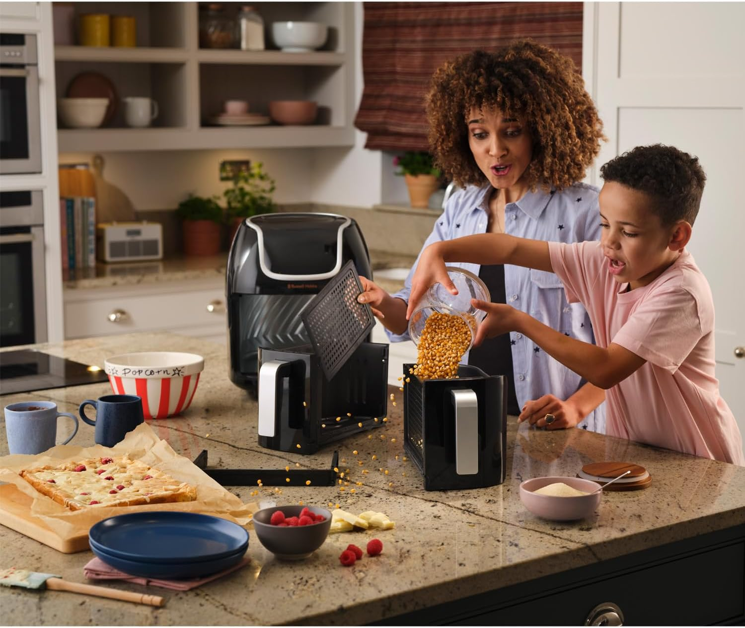 Kobieta i dziecko przygotowują popcorn w kuchni z frytownicą i różnymi produktami spożywczymi.