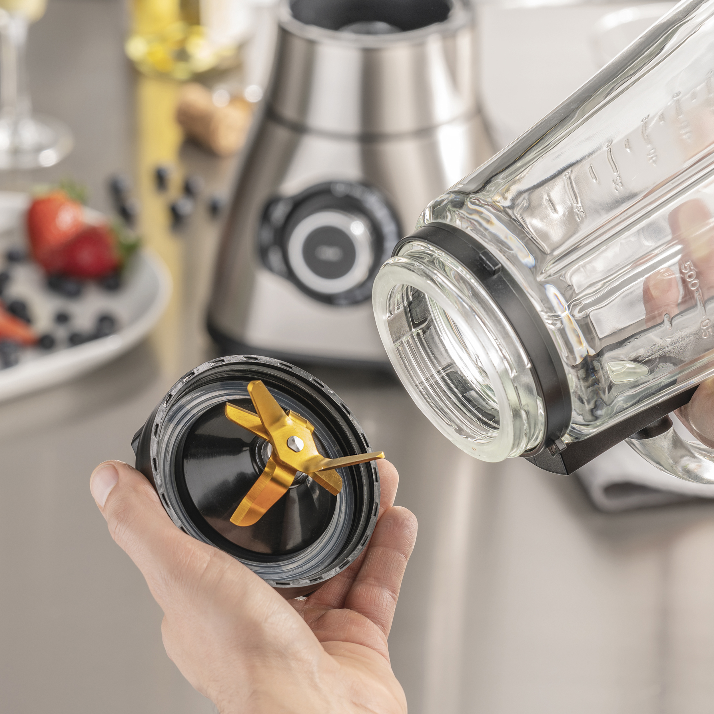 Die Hand einer Person, die einen Teil eines Mixers hält. Ein Mixer und Obst sind im Hintergrund.
