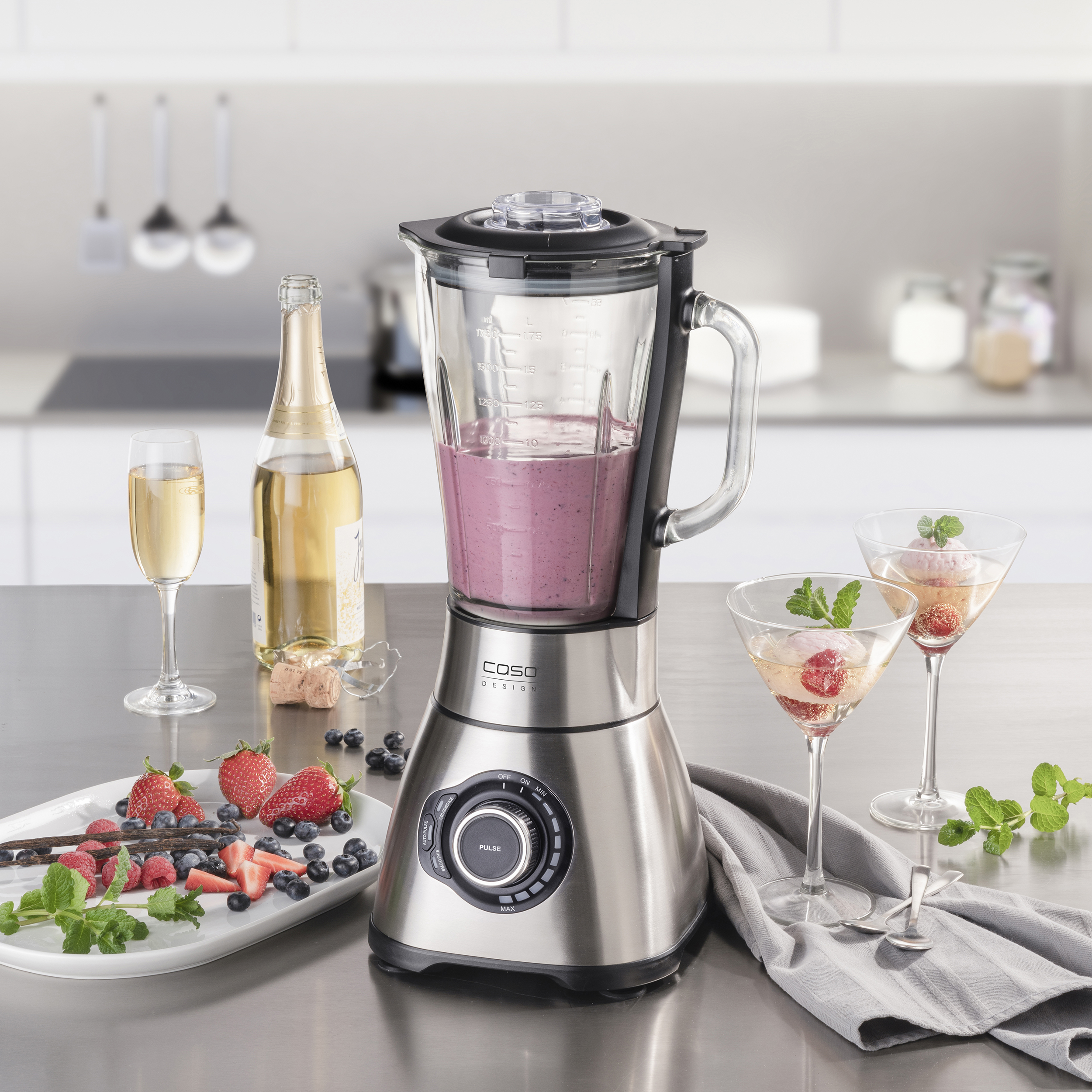 Ein Mixer mit einem rosa Smoothie, Zutaten und zwei Martinigläsern mit Beeren und Minze.