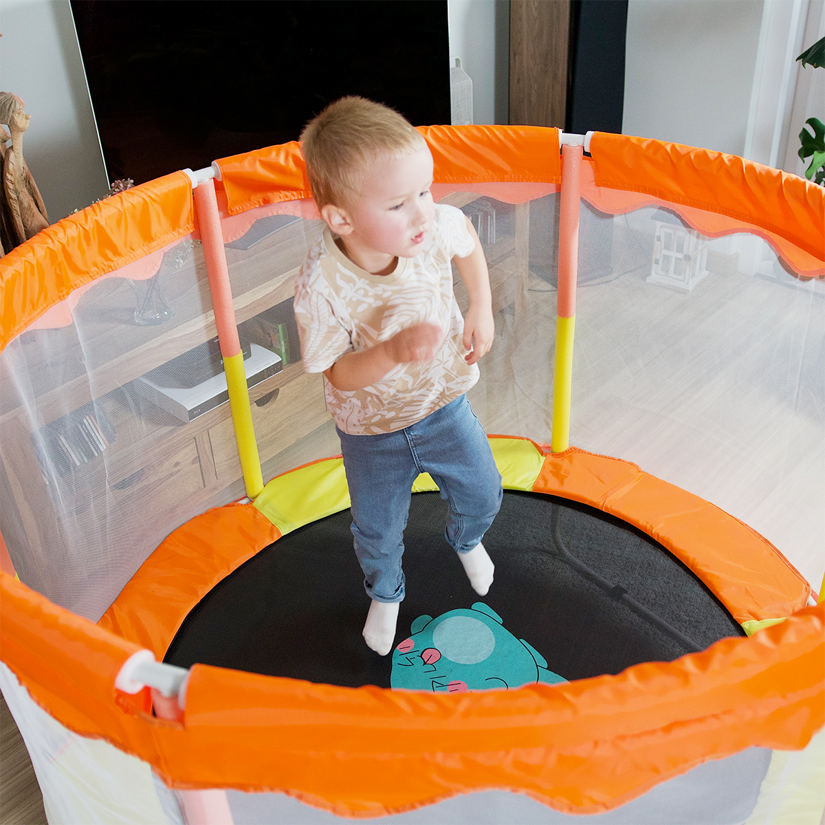 Junge springt in einem orangefarbenen und weißen Trampolin mit Sicherheitsnetz, in einem Raum. Blondes Haar, blaue Jeans.