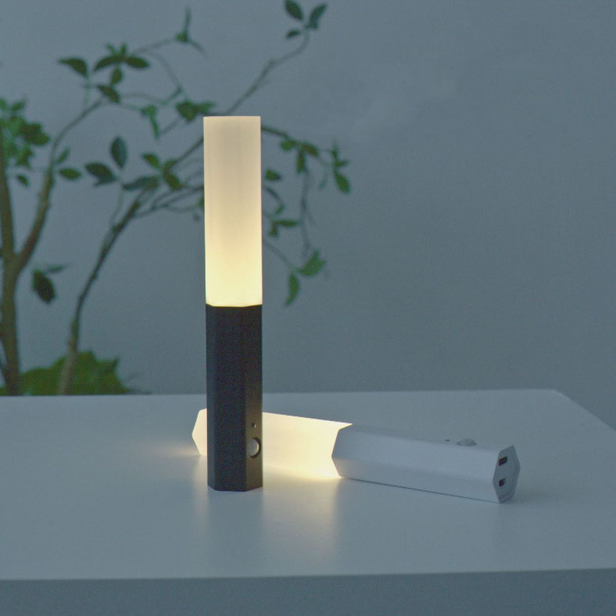 Zwei moderne Nachtlichter: eines aufrecht, eines horizontal. Beide haben einen weißen, beleuchteten oberen Teil und eine dunkle Basis.