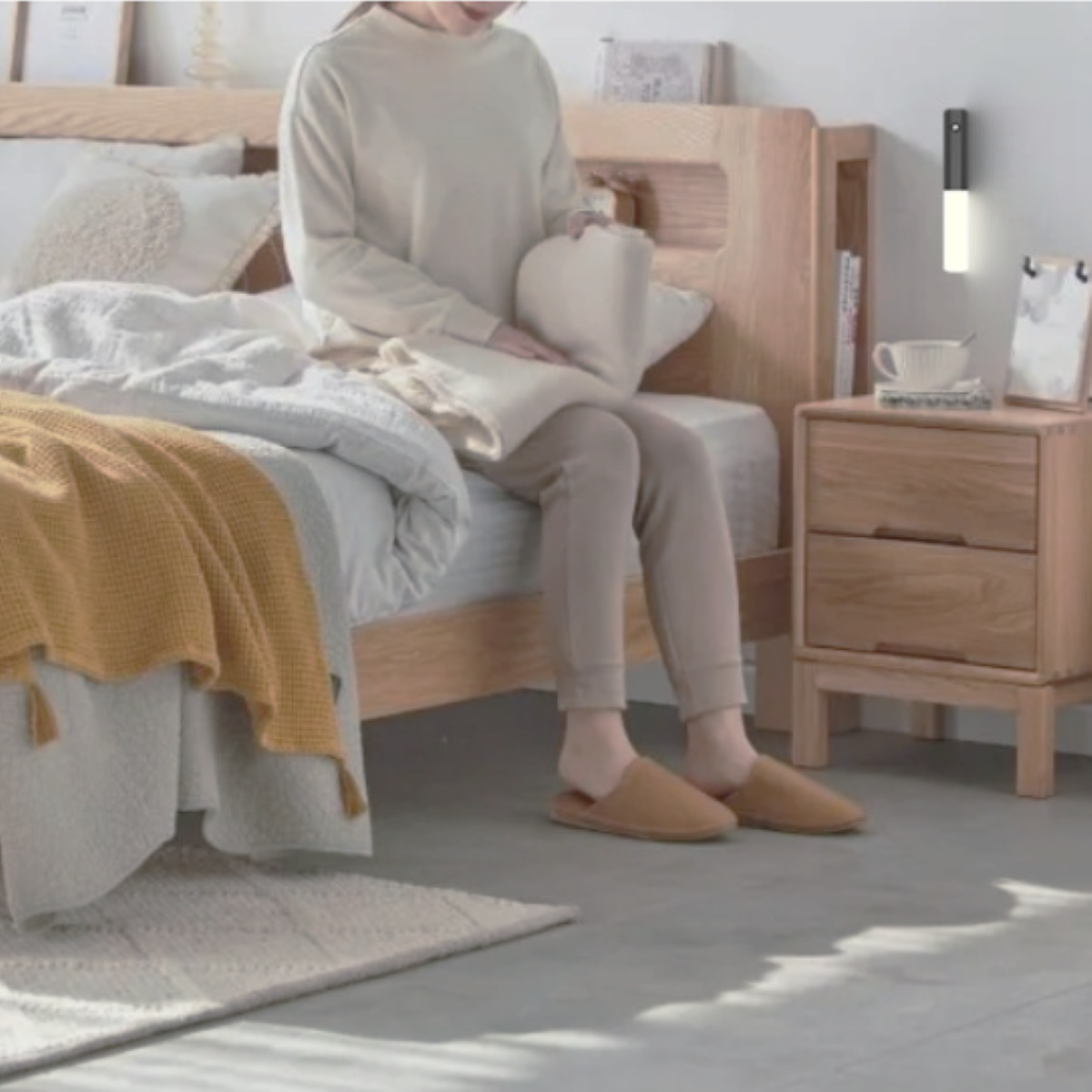 Eine Person sitzt auf einem Bett und hält eine Decke. Ein Nachttisch und eine Wandlampe sind sichtbar.