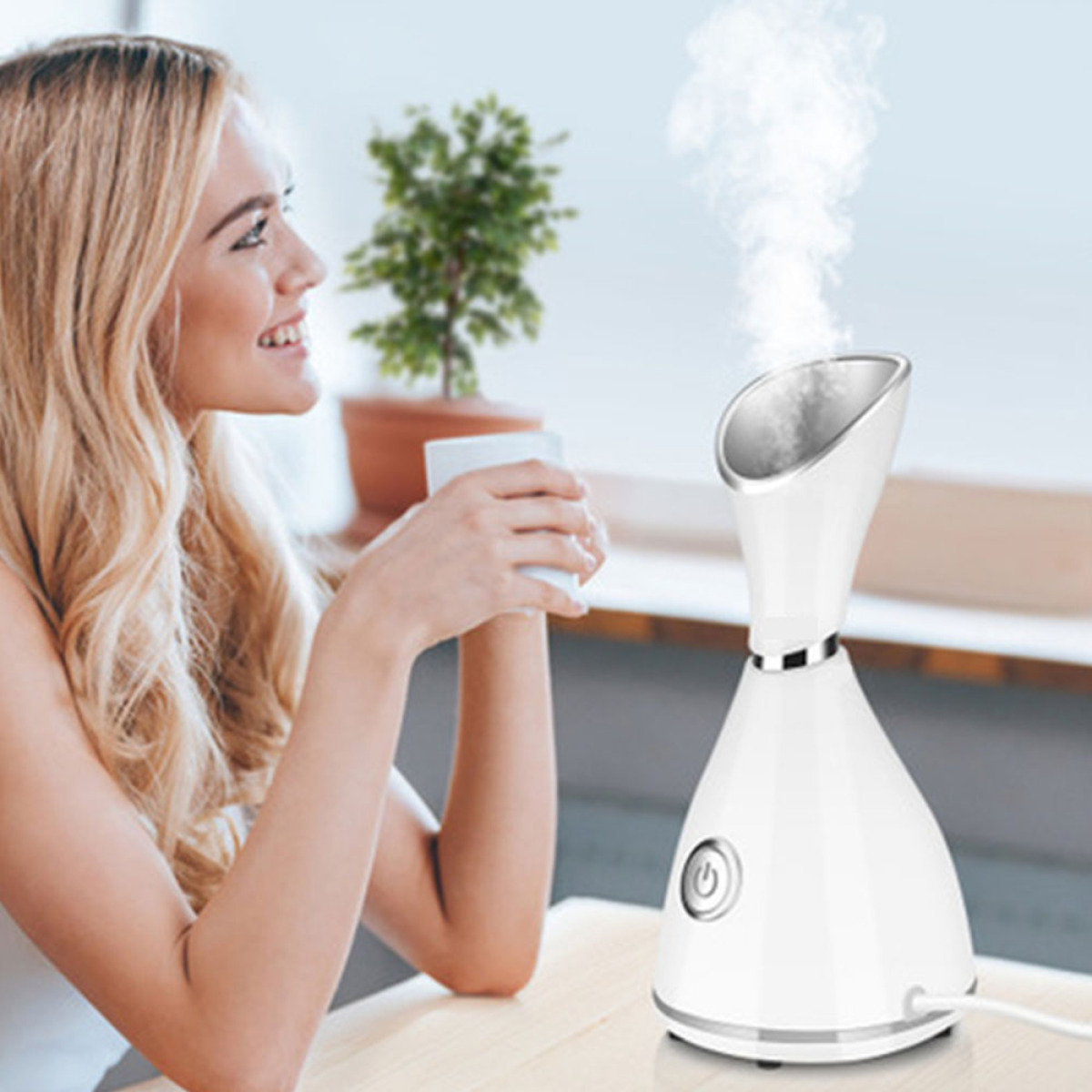 Frau hält eine Tasse, Dampf steigt aus einem weißen Gesichtsdampfer auf. Eine kleine Pflanze ist ebenfalls vorhanden.