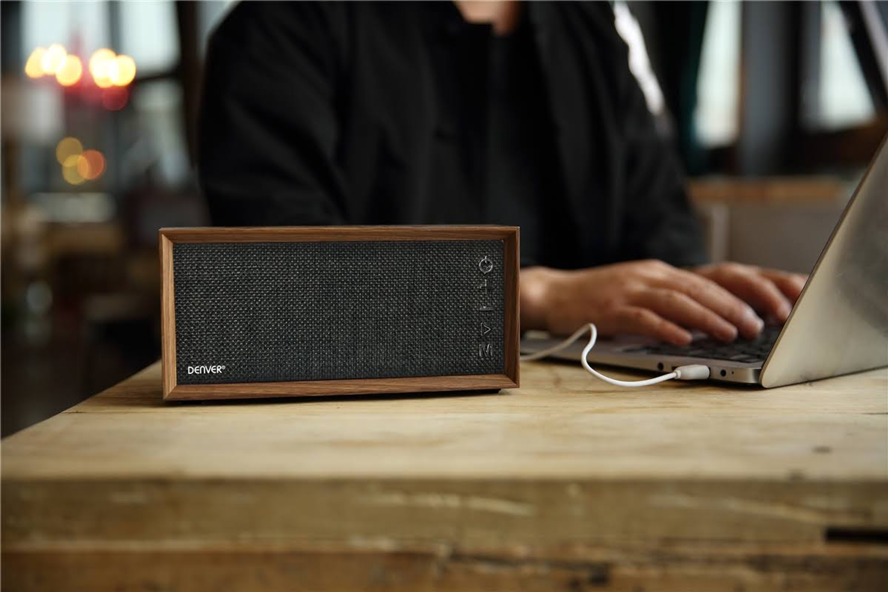 Un altavoz de madera con una pantalla de malla negra sobre una mesa de madera. Persona usa portátil.