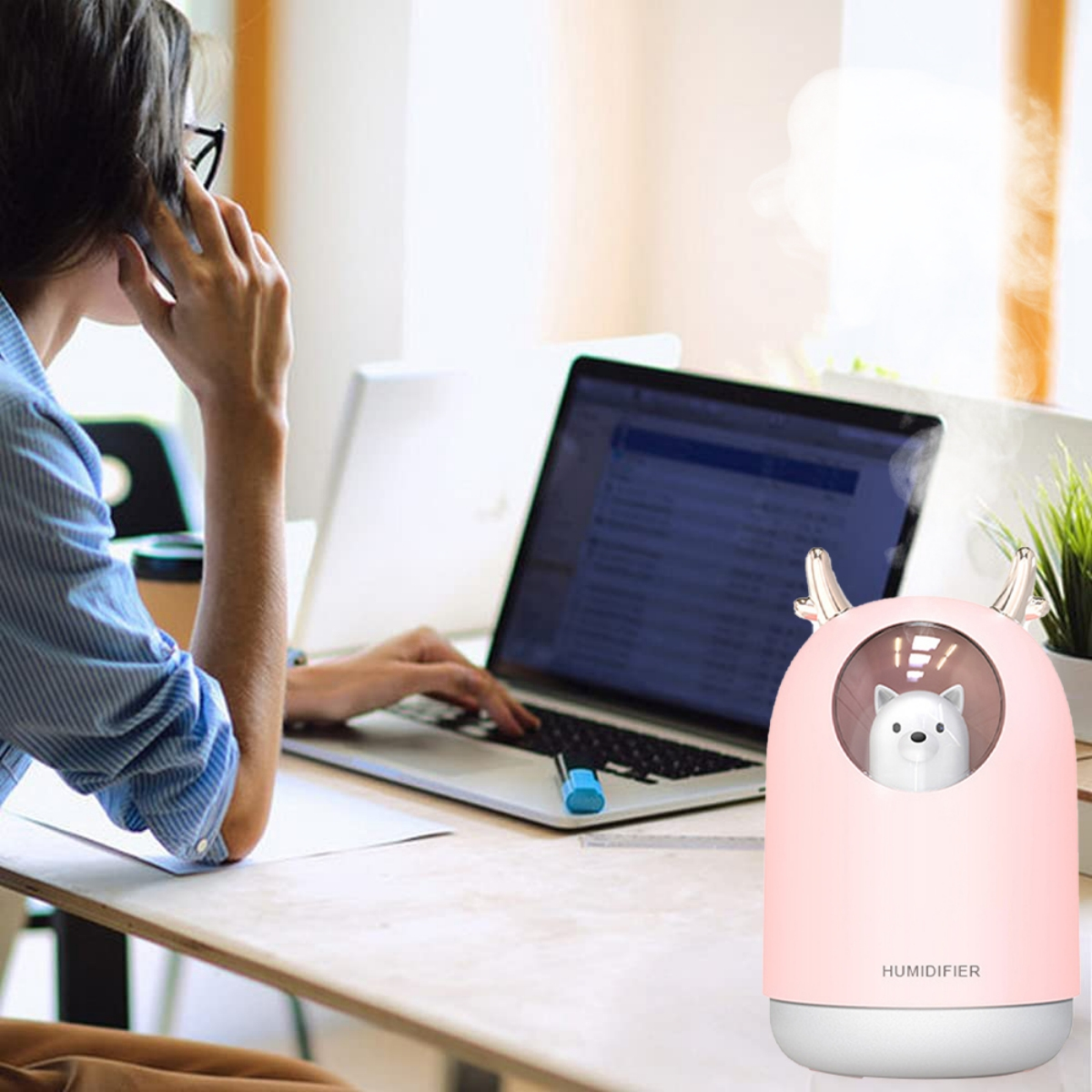 Frau telefoniert und arbeitet am Laptop. Ein rosa Luftbefeuchter mit Bär steht auf dem Tisch.