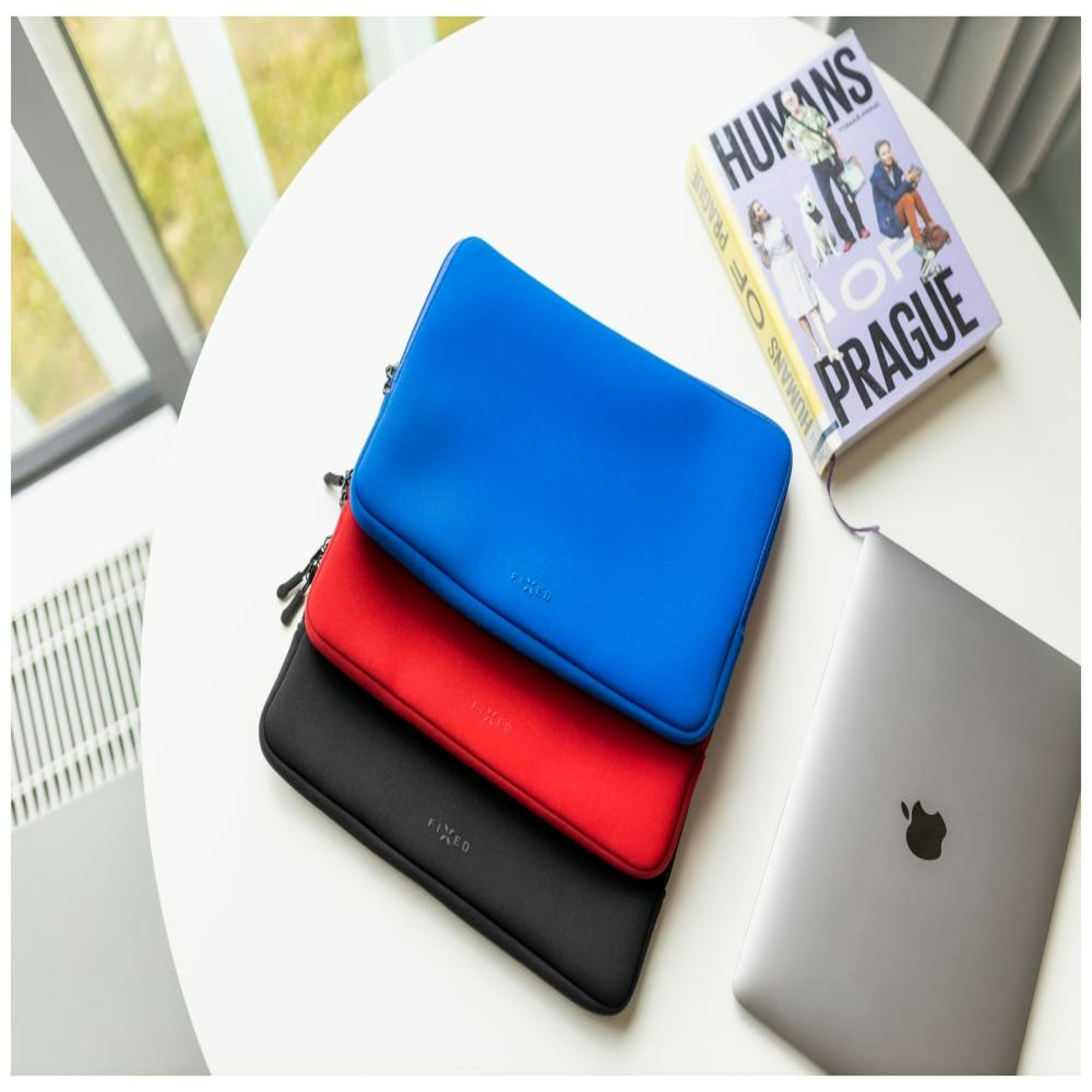 Drei Laptop-Hüllen in Blau, Rot und Schwarz auf weißem Tisch, mit Buch und Laptop.