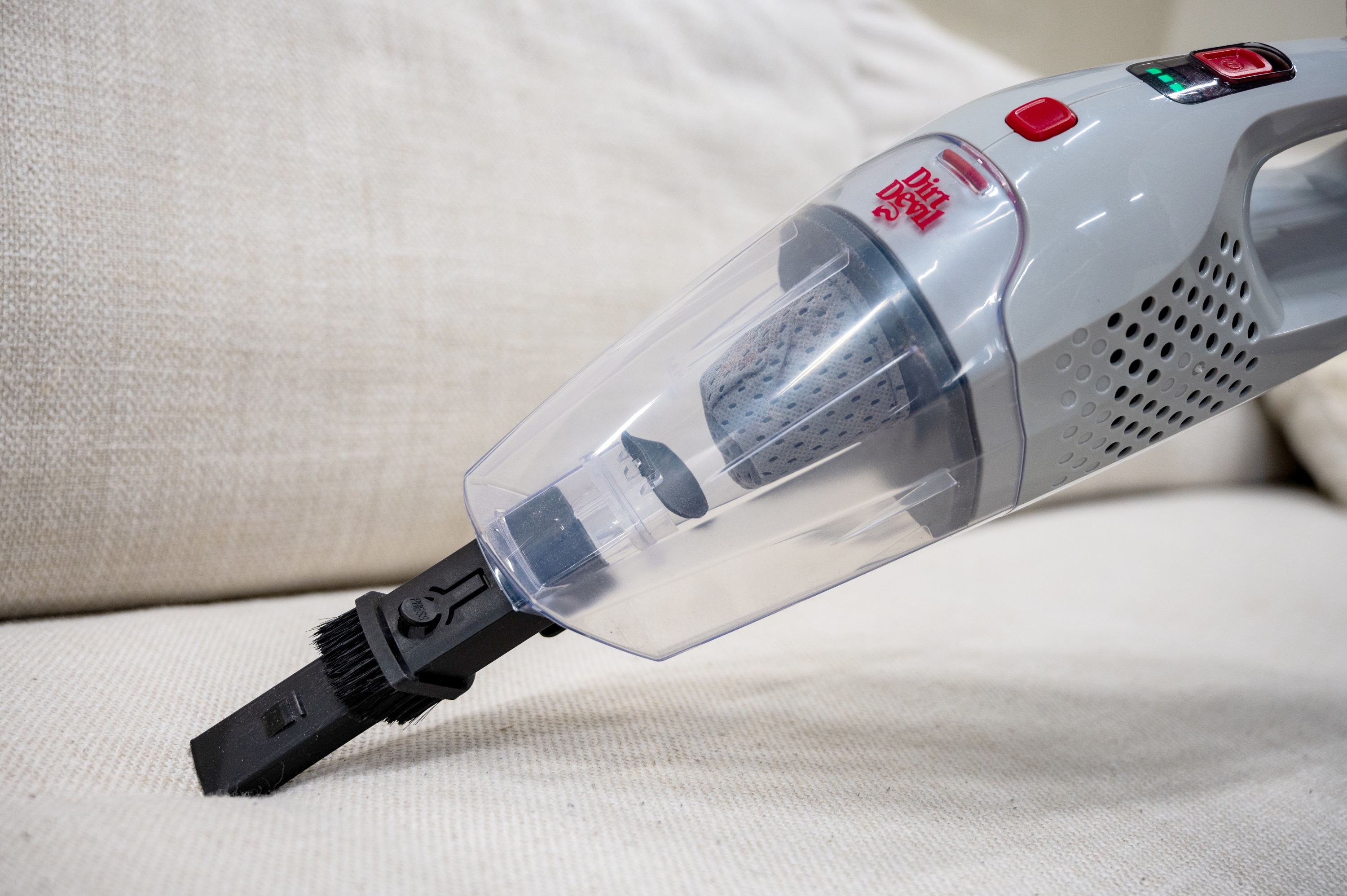 Ein grau-roter Dirt Devil Staubsauger, der ein cremefarbenes Sofa reinigt.
