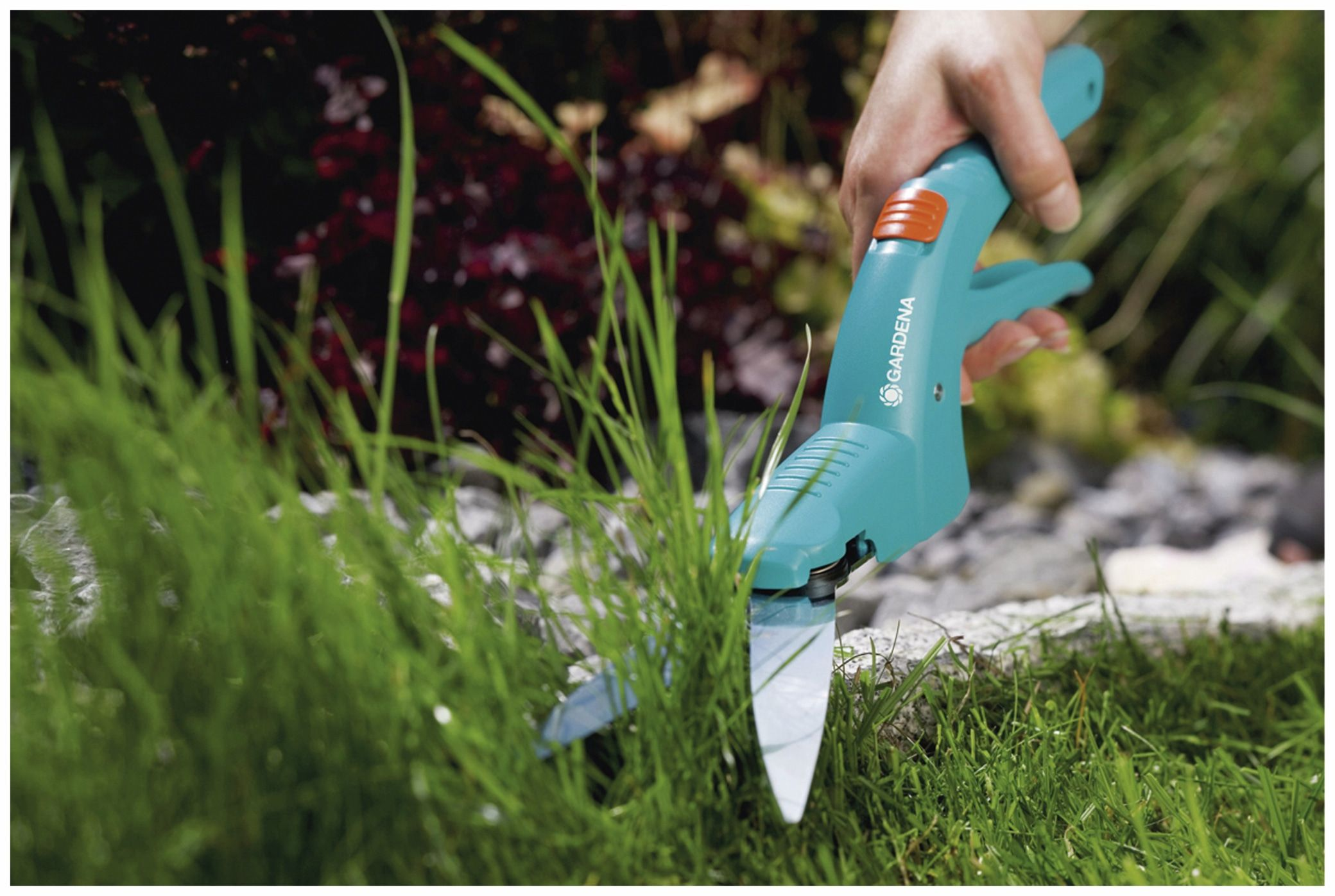 Ręka trzyma turkusowe nożyce Gardena, które tną zieloną trawę. Tło pokazuje ogród z zieloną trawą i ciemnymi kwiatami.