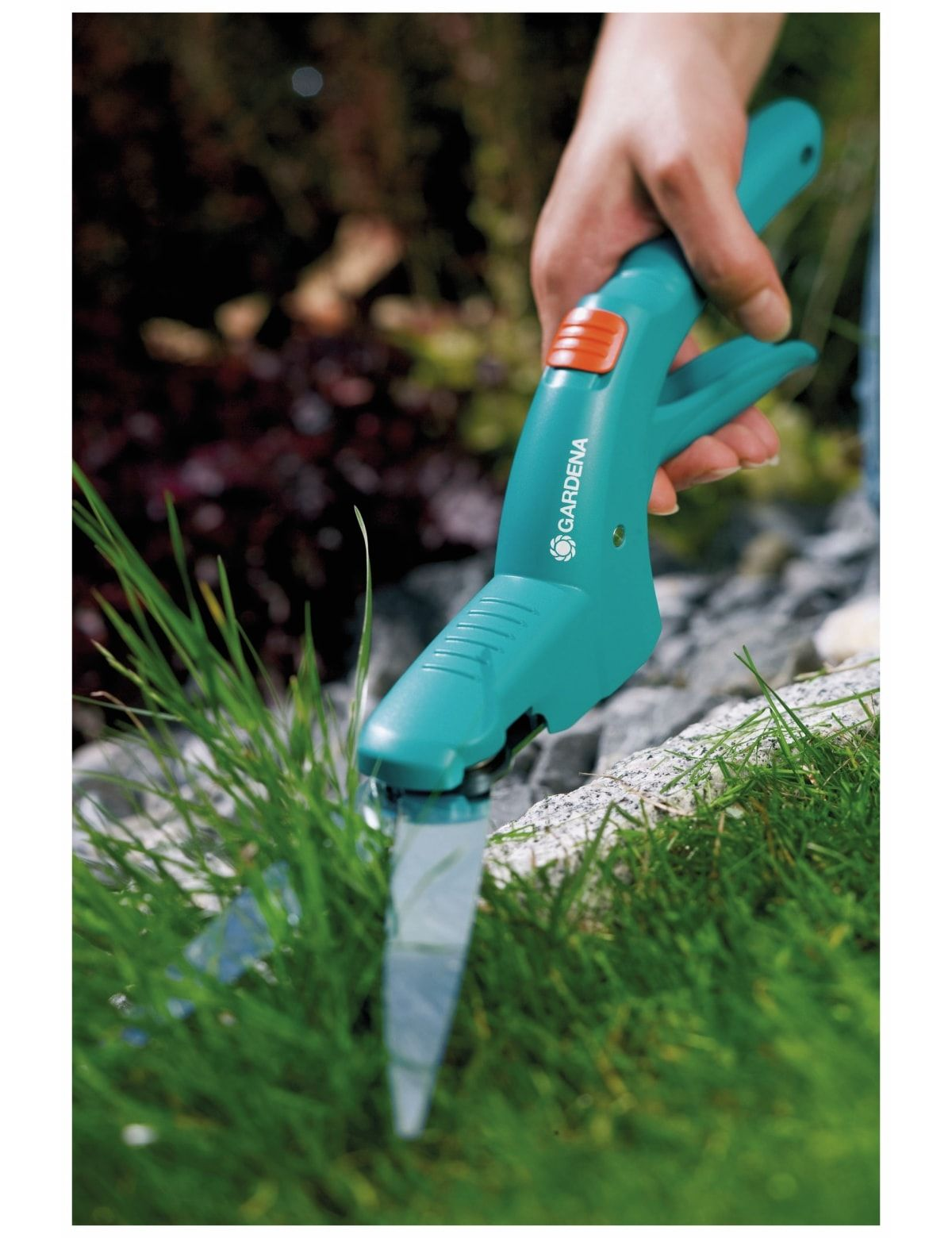 Ręka trzymająca turkusowe nożyce do trawy GARDENA, tnąca trawę. Tło stanowią ciemnozielone krzewy i jasnozielona trawa.