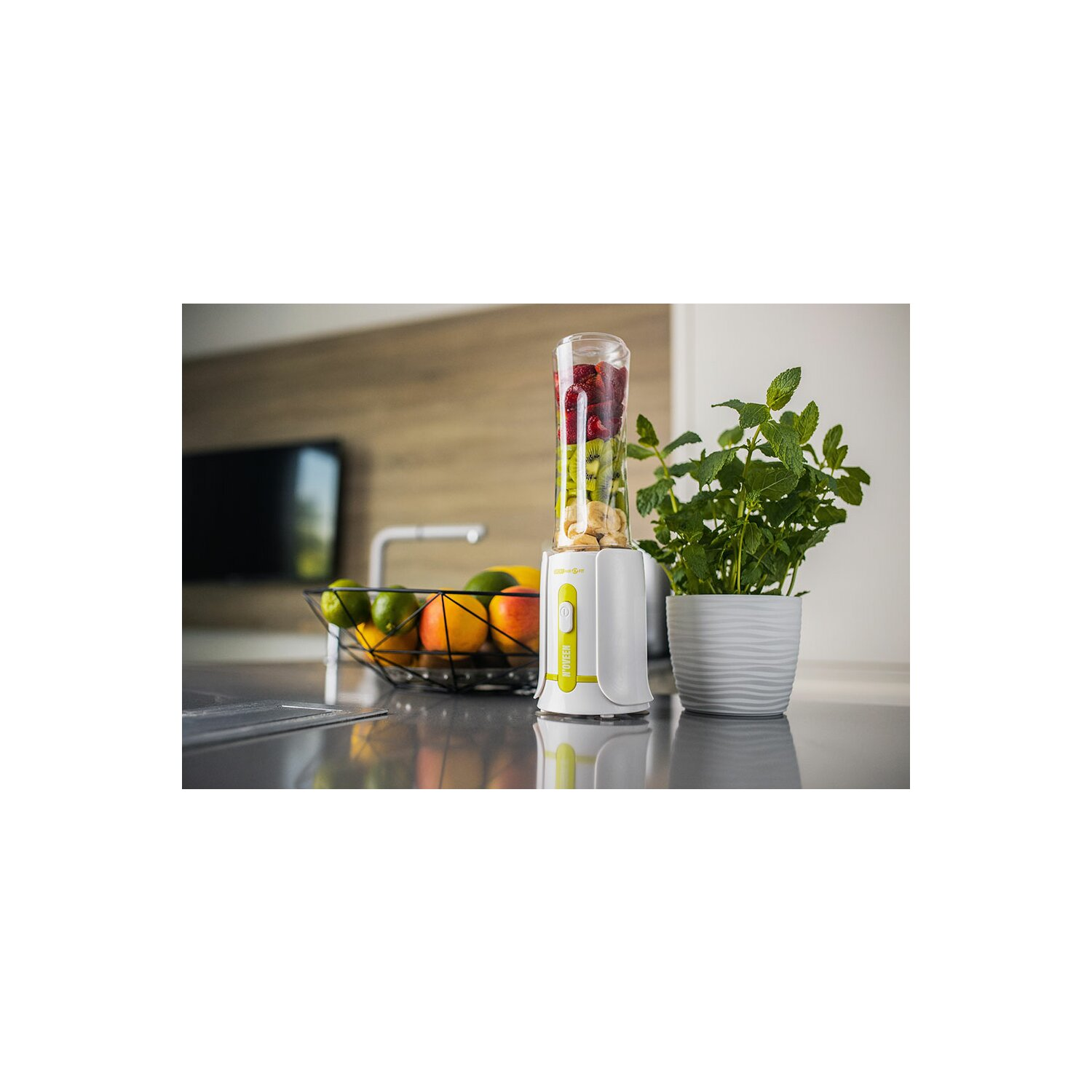 Biały i zielony blender z owocami w środku, na blacie kuchennym w pobliżu rośliny.