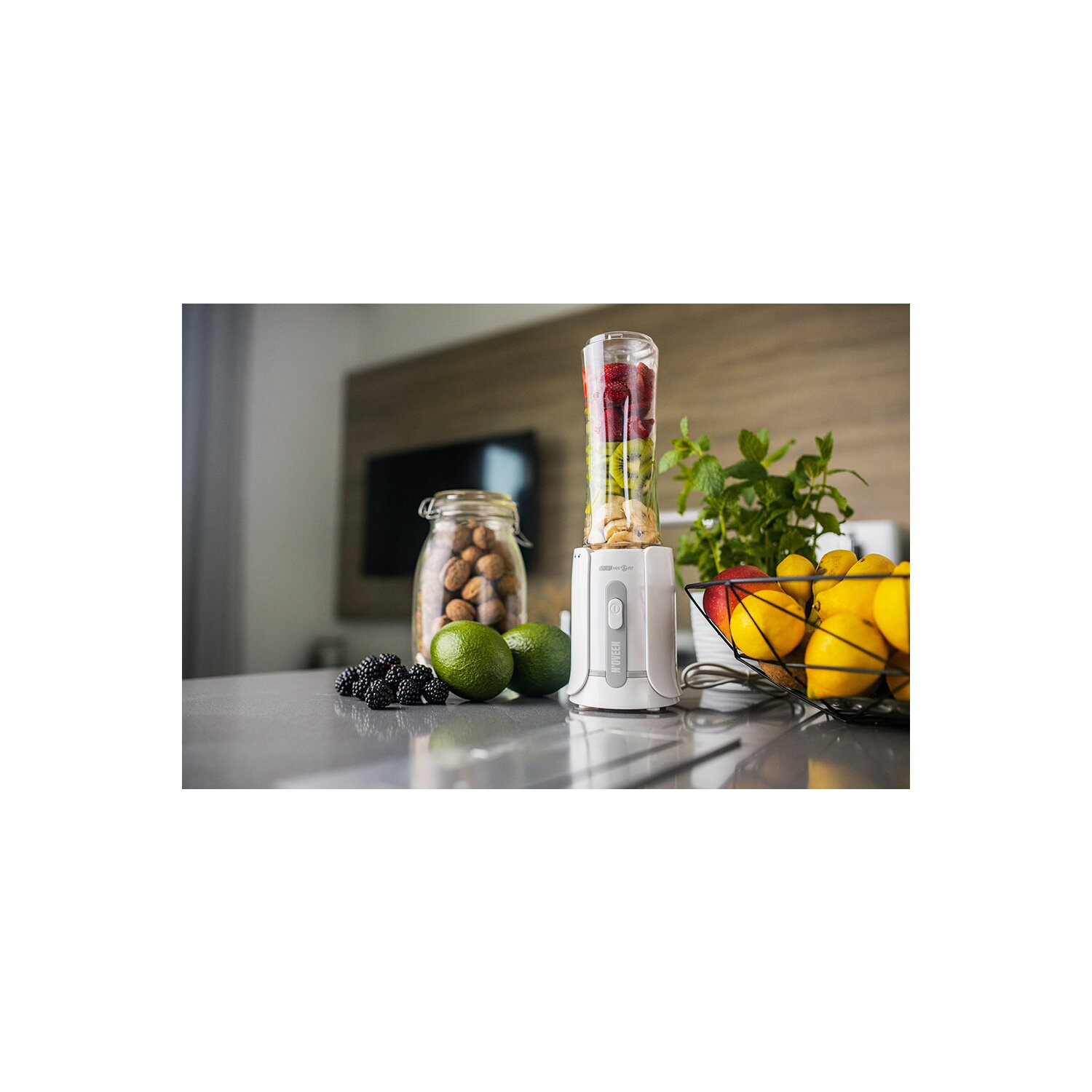 Biały blender z owocami, limonkami, jeżynami i cytrynami w drucianym koszu na szarym blacie.