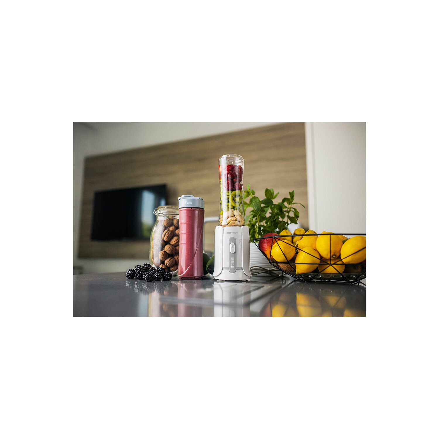 Blender z owocami w środku, słoik orzechów i miska cytryn na blacie kuchennym.