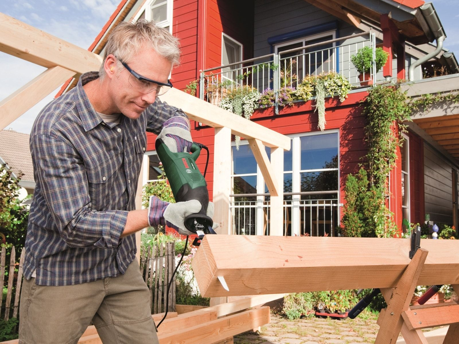 Mężczyzna używa piły. Tnie drewno na kozłach. Dom w tle.
