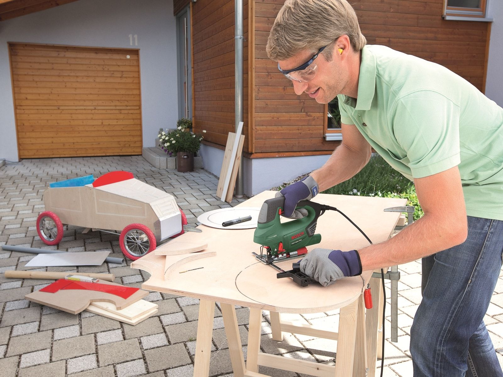 Mężczyzna używający wyrzynarki na drewnianej powierzchni, obok zabawkowy samochód i kawałki drewna.