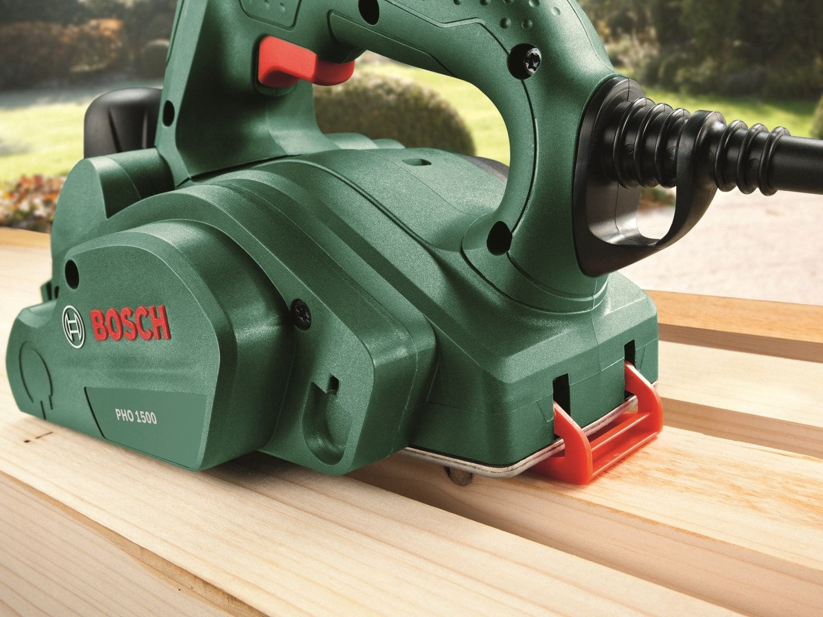 Un homme rabote une planche de bois avec un rabot Bosch sur un établi dans un jardin.