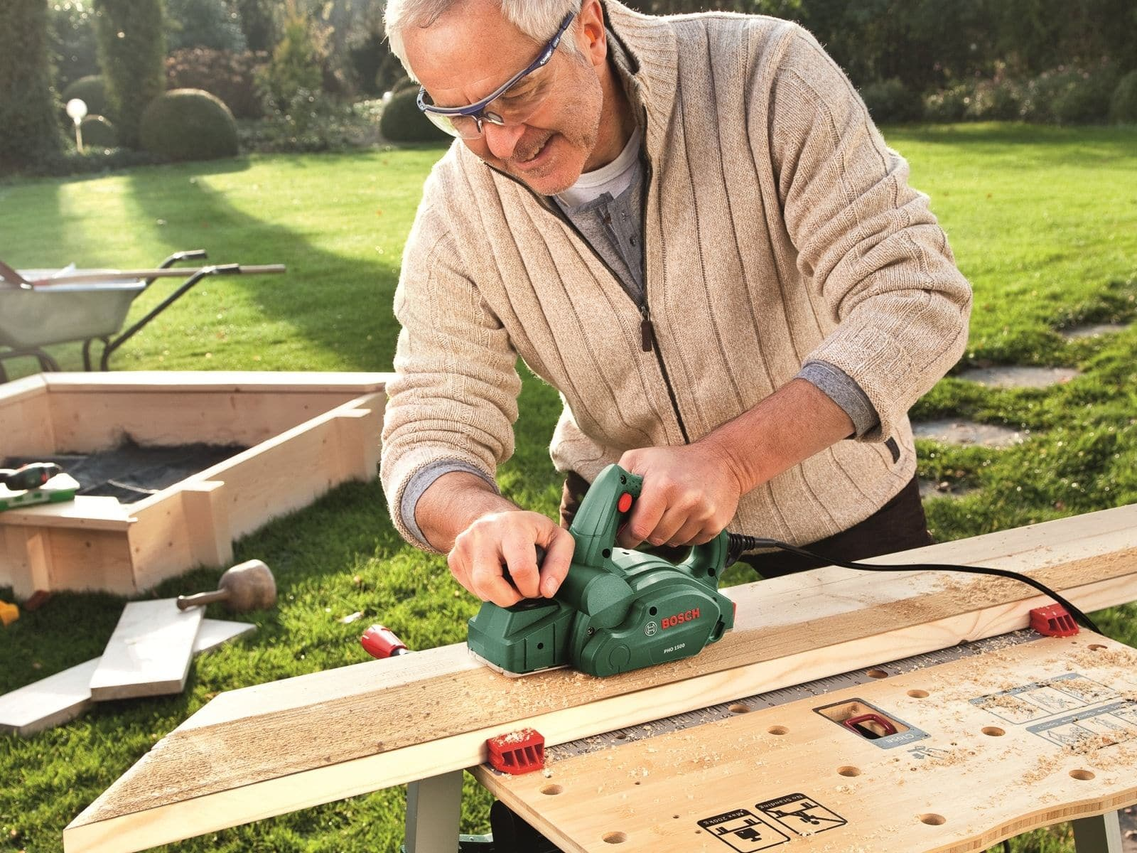 Un homme rabote une planche de bois avec un rabot Bosch vert sur un établi dans un jardin.