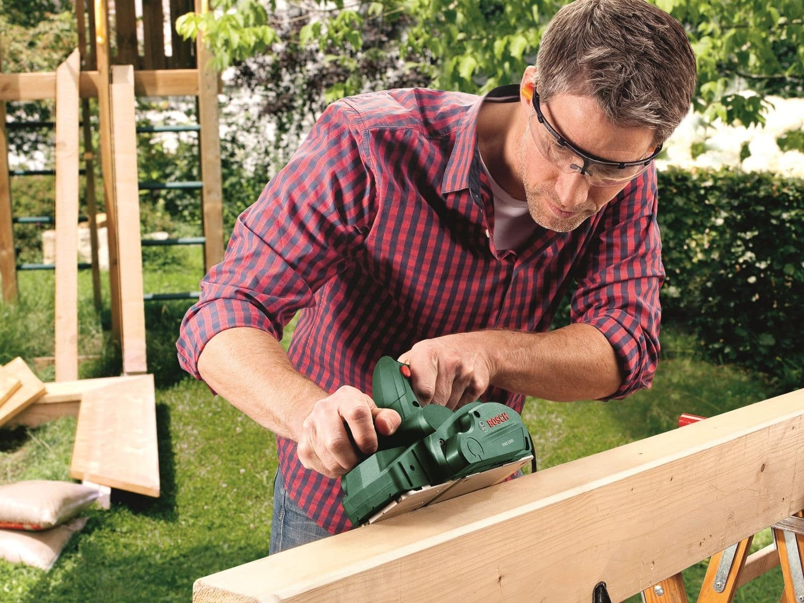 Un homme utilise un rabot électrique Bosch vert sur une planche de bois dans un environnement de jardin.