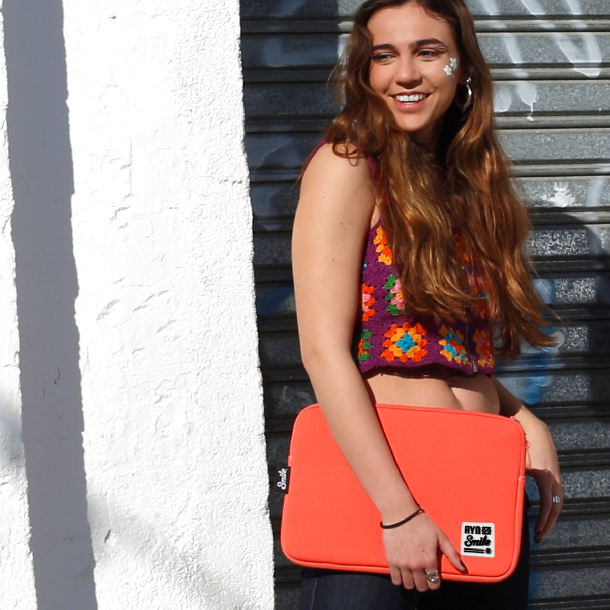 Donna con top all'uncinetto, che tiene una custodia arancione per laptop. Sorride davanti a un muro.