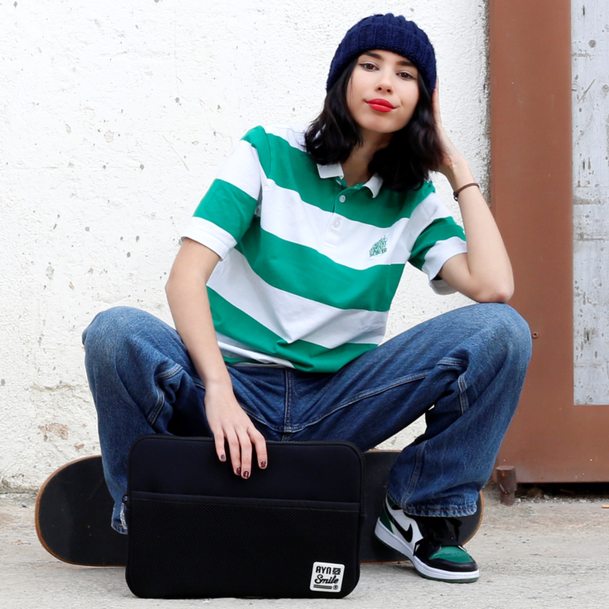 Giovane donna con berretto e skateboard tiene una custodia nera per laptop. Indossa una maglietta a righe.