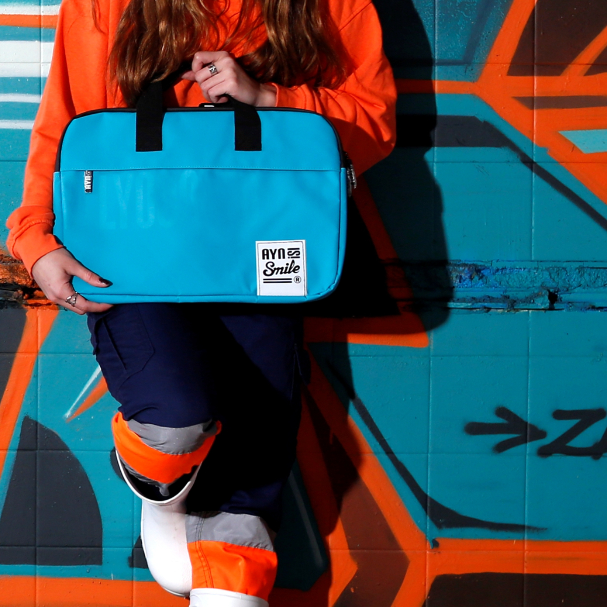 Persona che tiene una borsa blu per laptop con un logo, di fronte a un muro di graffiti.
