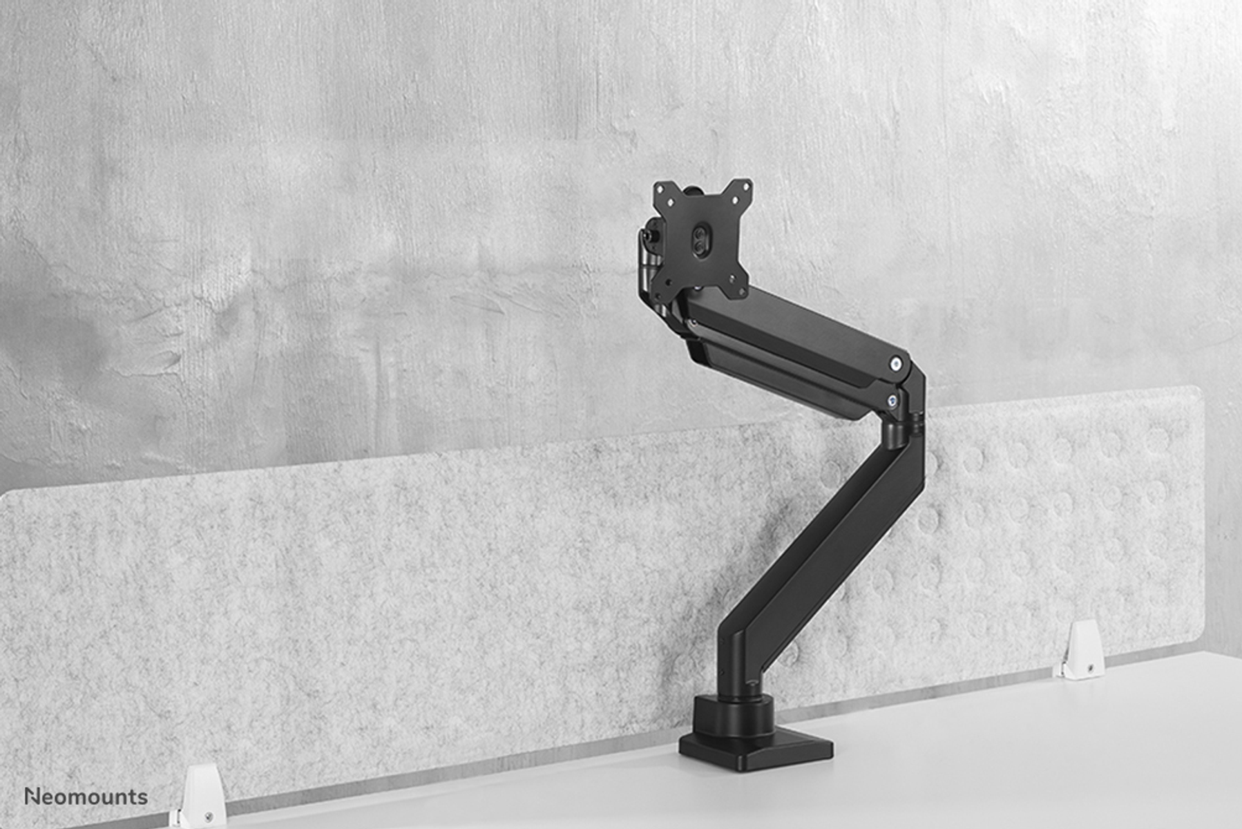 A black monitor arm with a flexible mount stands on a desk in front of a wall.