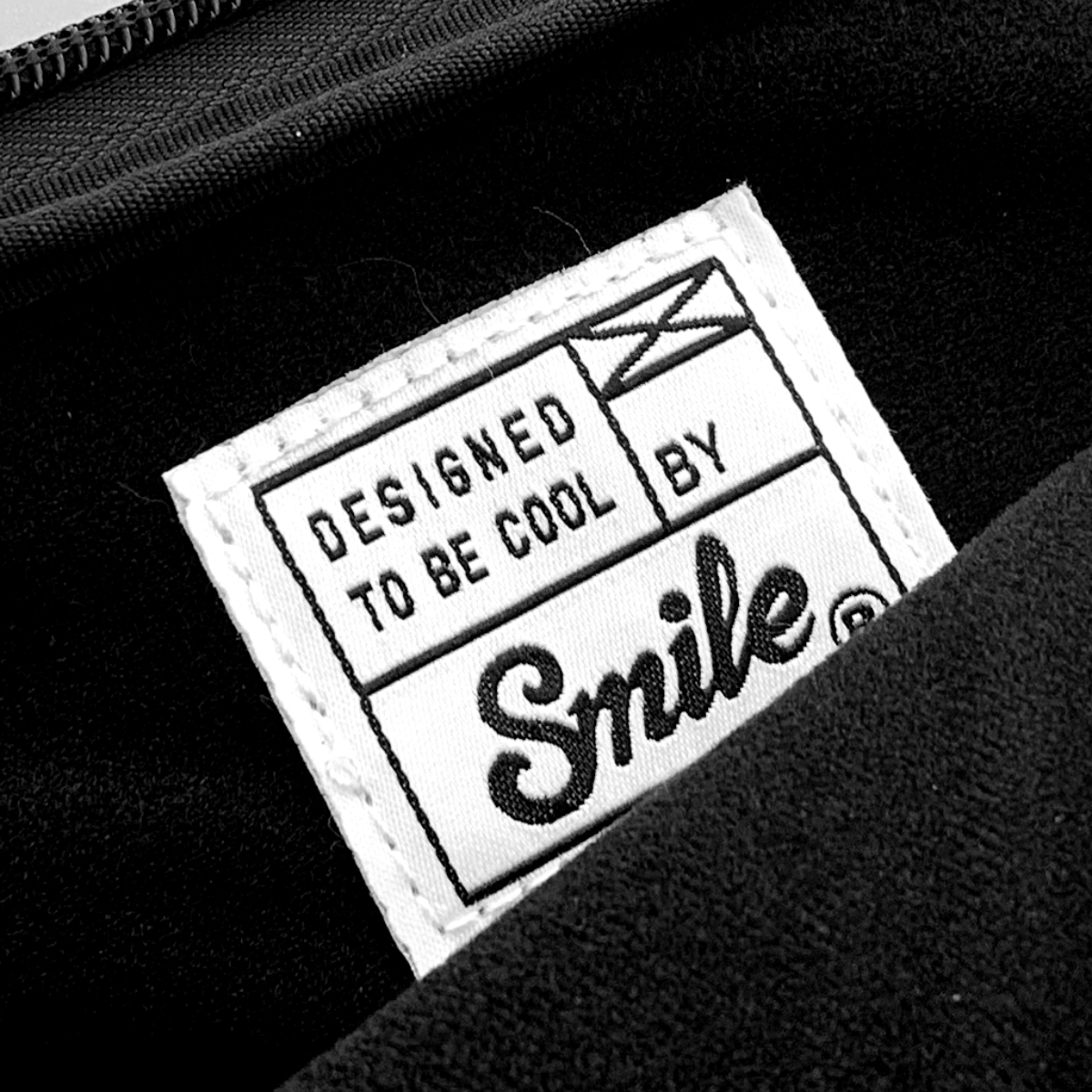 Primer plano de una etiqueta blanca sobre tela negra. La etiqueta dice 'DESIGNED TO BE COOL BY Smile'.