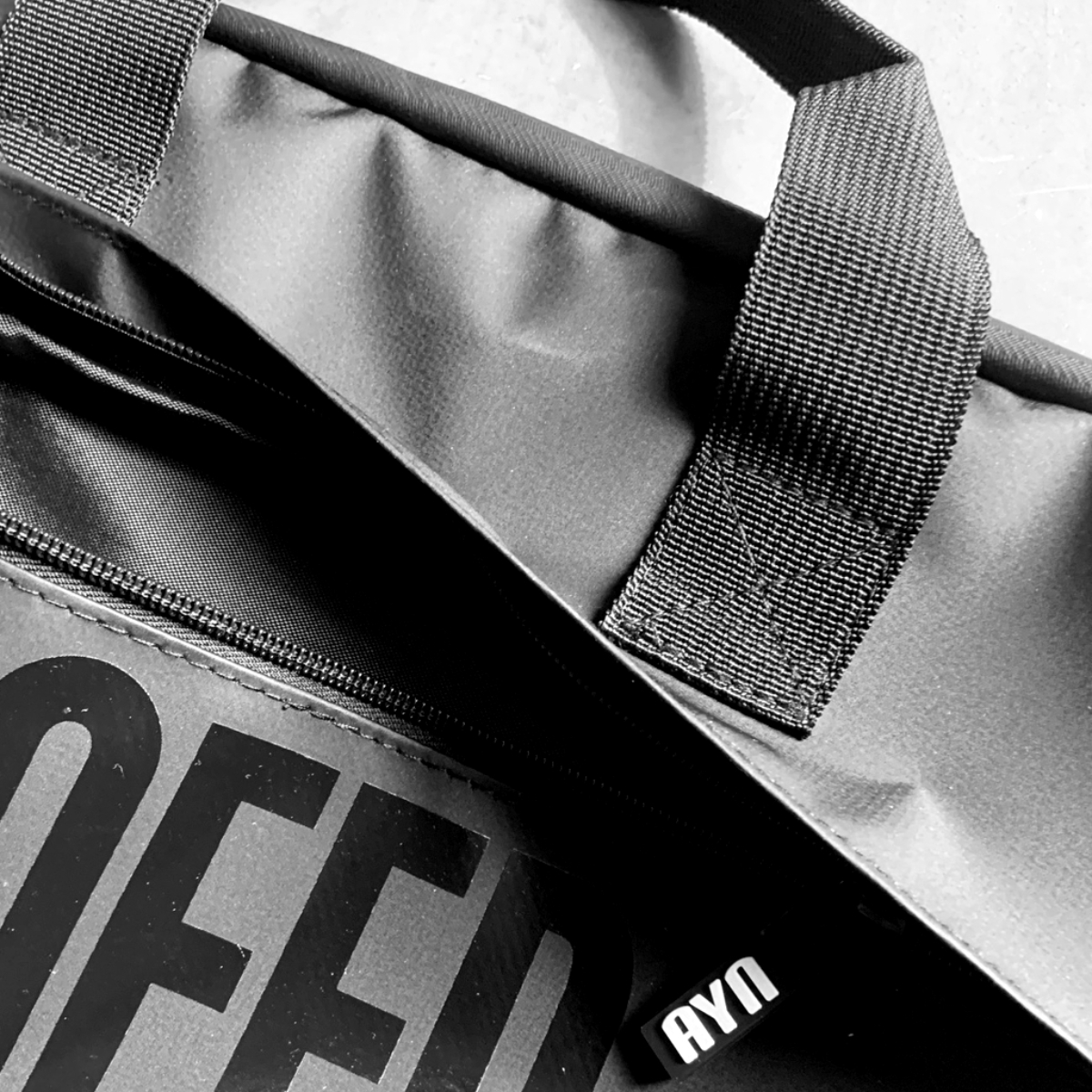 Bolso negro con texto OFF grande y etiqueta AYN. Vista en ángulo sobre una superficie.