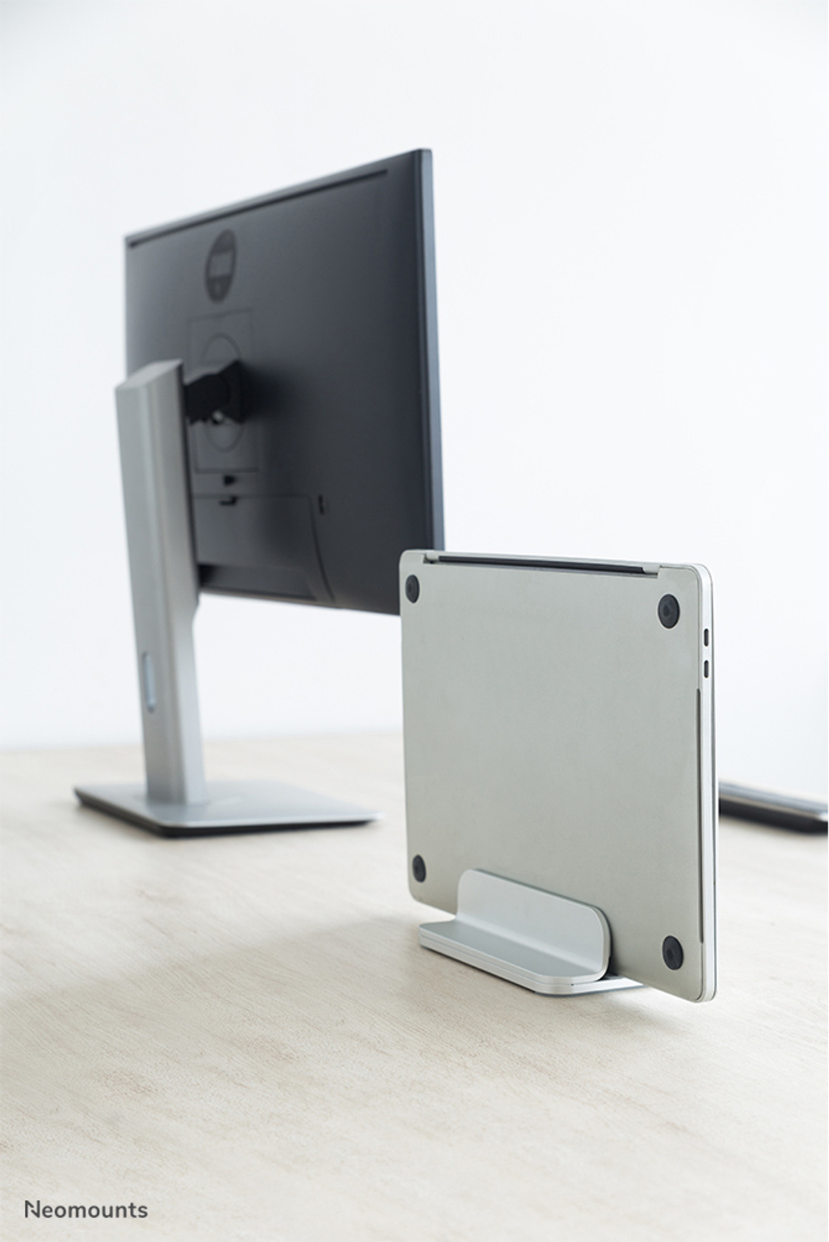 A laptop stands on a slim stand in front of a computer monitor.